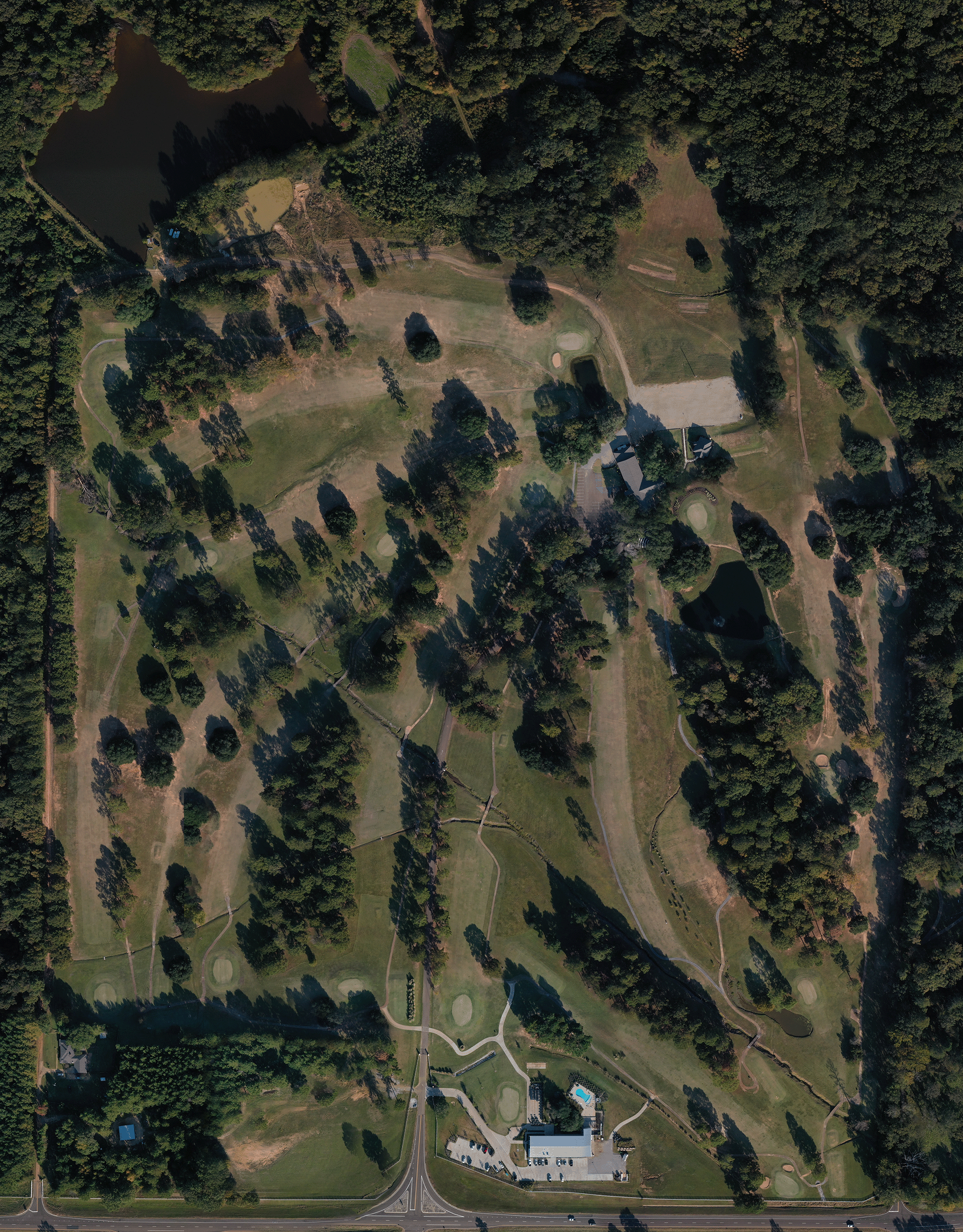Aerial view of a golf course surrounded by trees and a pond, with pathways, sand traps, and a clubhouse at the bottom.