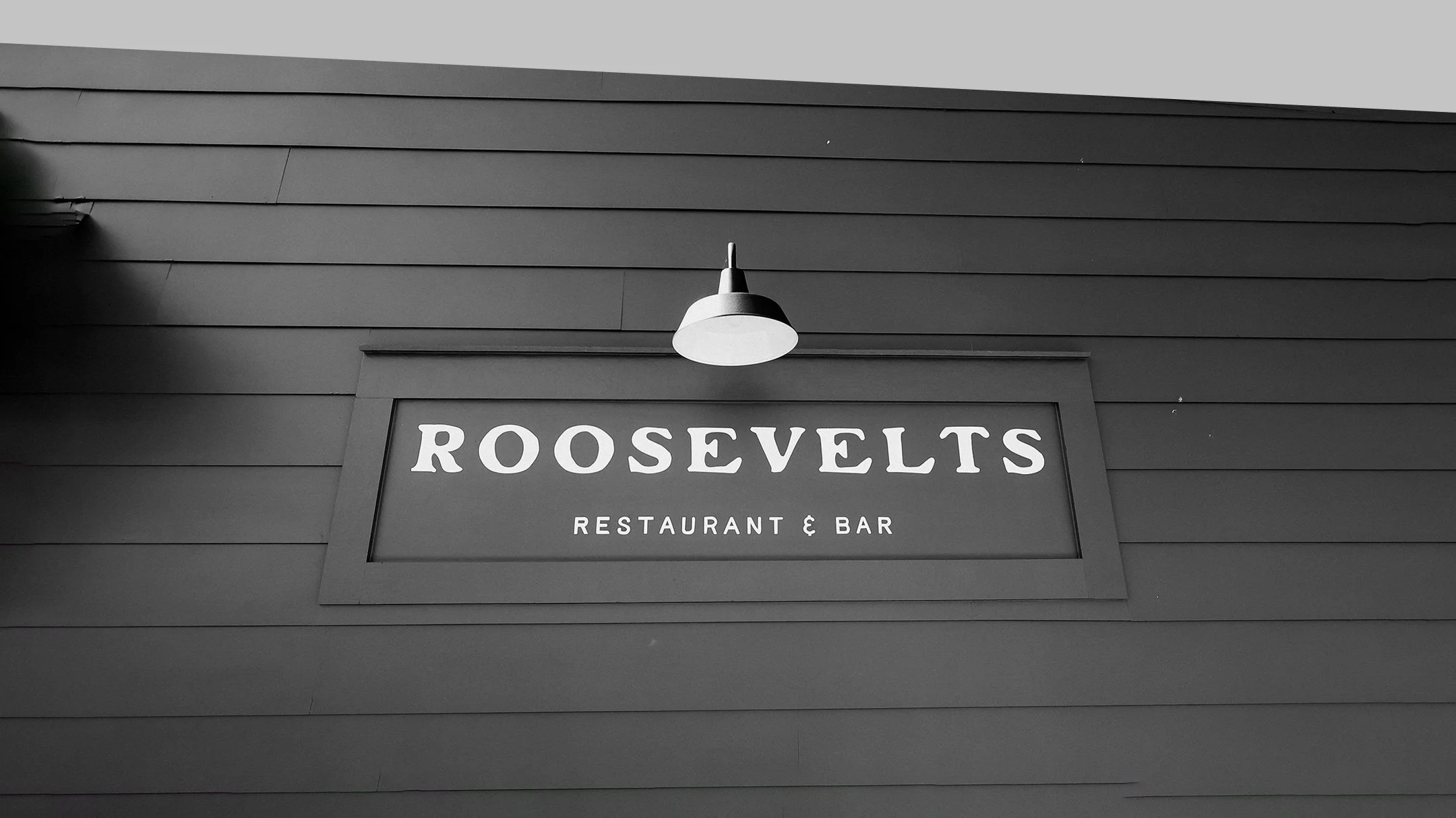 Exterior sign for Roosevelts Restaurant & Bar at Live Oaks Golf Club in Jackson, Mississippi, featuring white serif lettering on a dark wood shiplap wall with a vintage gooseneck barn light above the sign.
