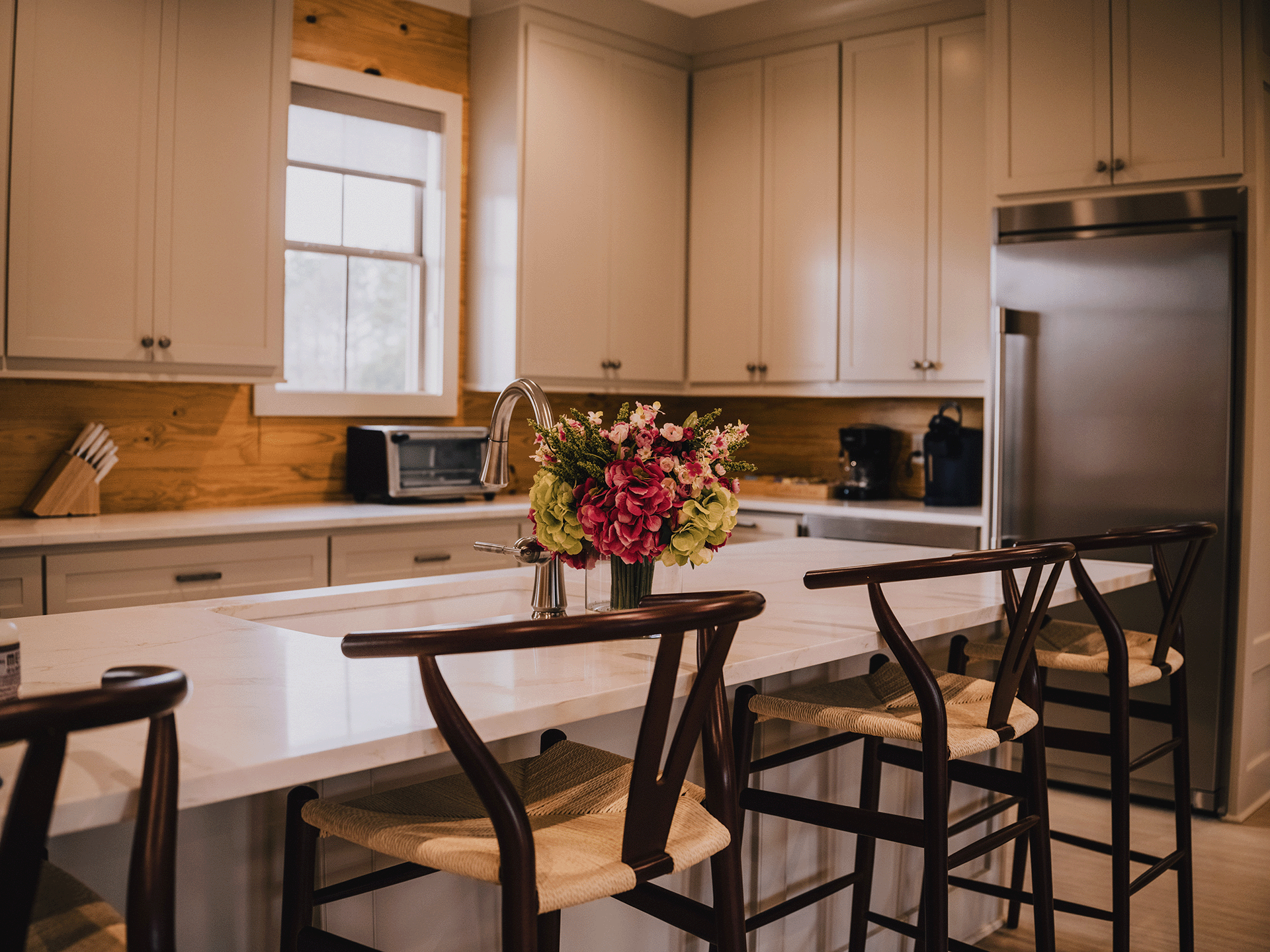 A modern kitchen with a white island counter, a vase of pink and green flowers, and dark wooden bar stools with woven seats. The kitchen has white cabinets, a window, a stainless steel refrigerator, and small appliances on the counter.