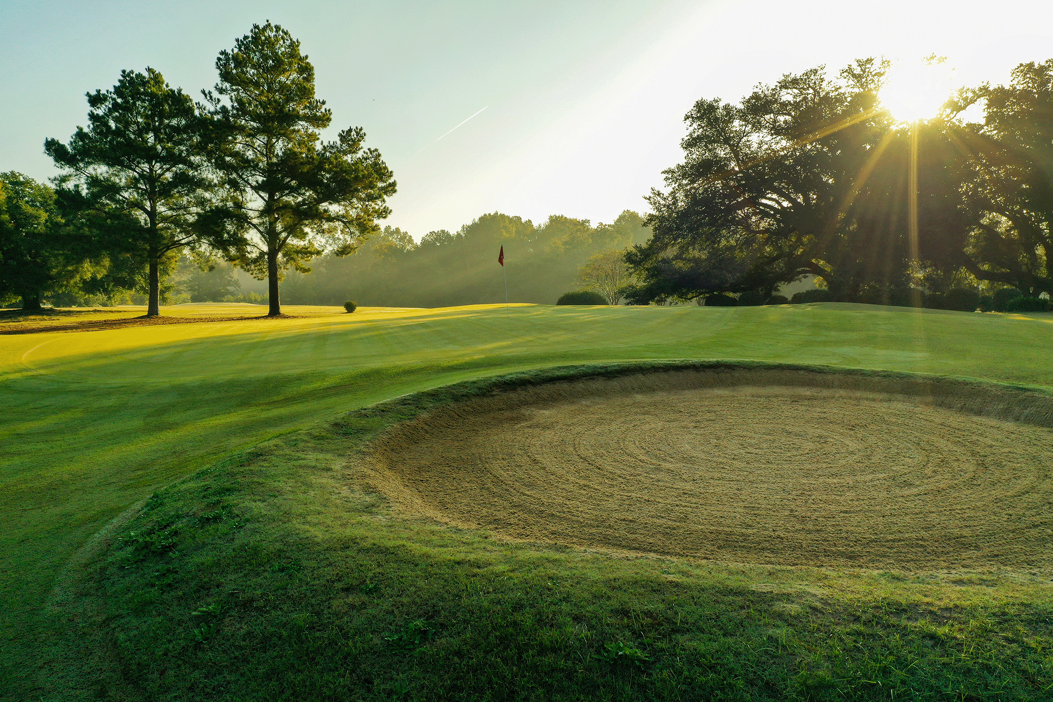 A 18 hole golf course green with a sand trap in the foreground, a flag on the green, and trees in the background, with sunlight filtering through on perfect greens next to a well maintained hazard.