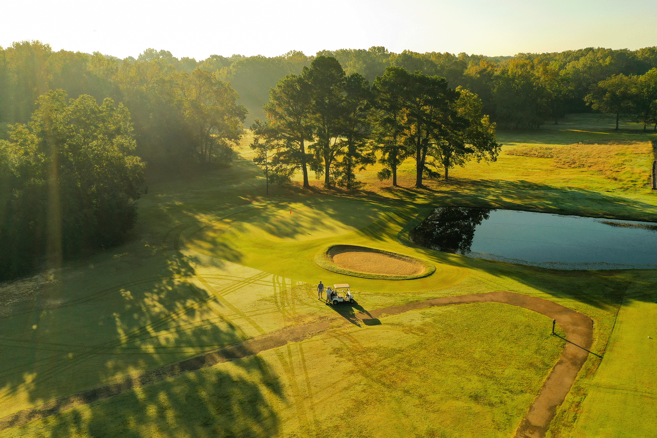 Aerial view of a golf course with a couple of people near a golf cart, surrounded by green grass, trees, a pond, and a sand bunker, during sunrise or sunset.