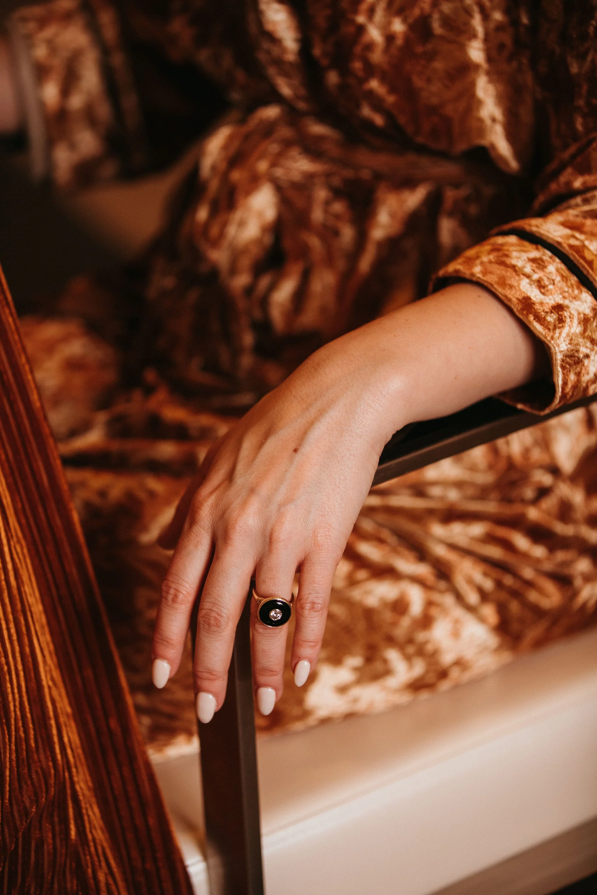 A woman's hand with a large dark ring resting on a metal bed frame, wearing a velvety patterned brown and gold robe.