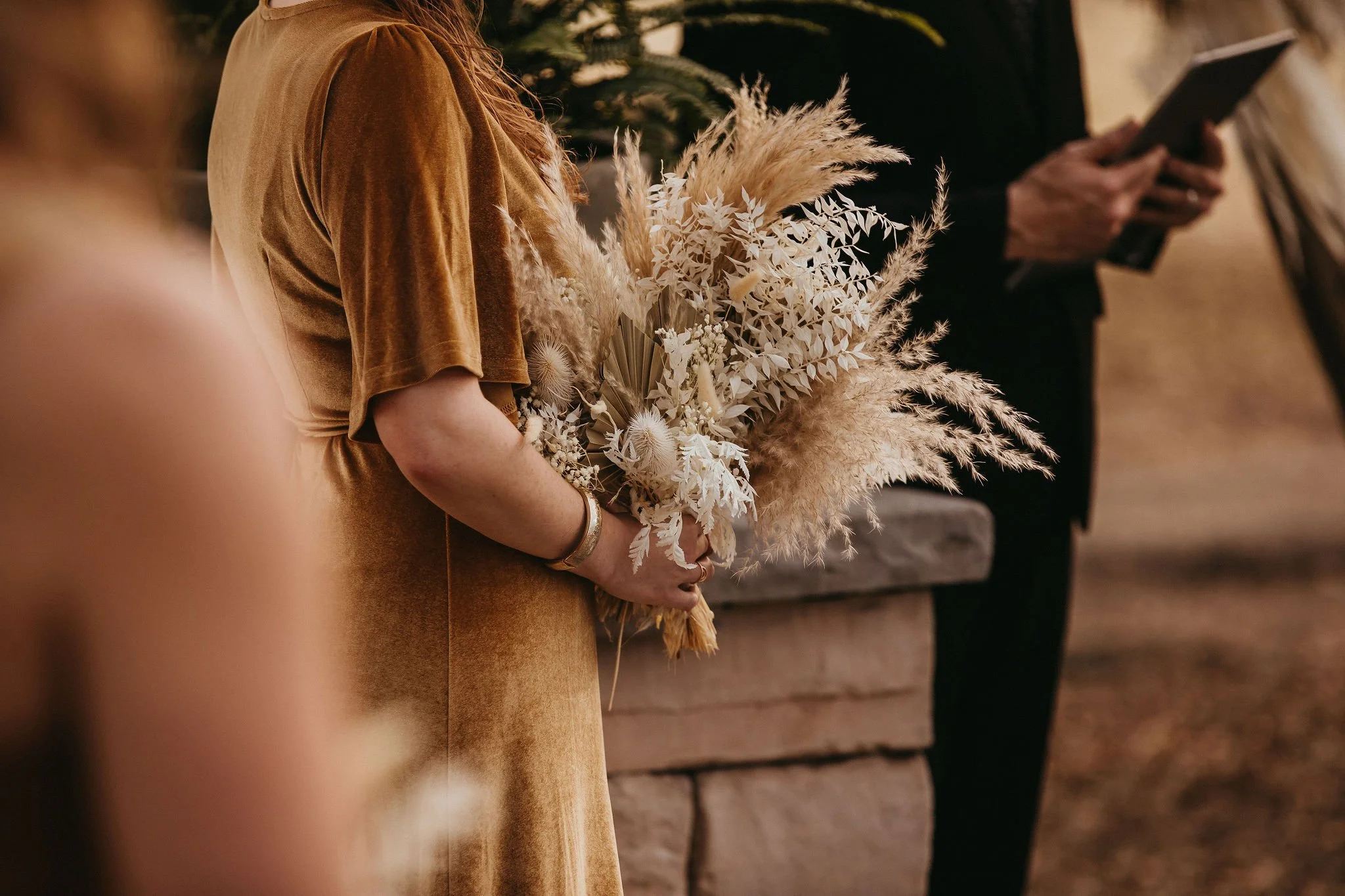 A woman in a brown velvet dress is holding a bouquet of dried flowers and grasses, standing in a warmly lit indoor setting.