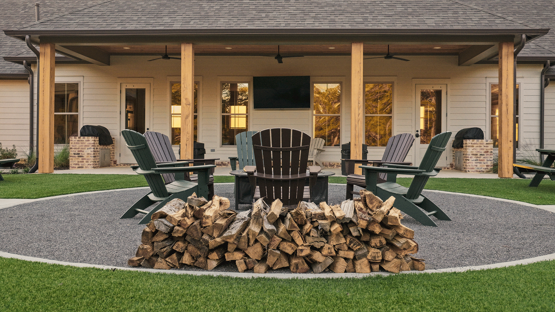 Backyard patio with six Adirondack chairs directed towards a firewood pile in the center, surrounded by a gravel area, with a large house in the background featuring a screened porch, windows, and a mounted TV, with grass and landscaping around.