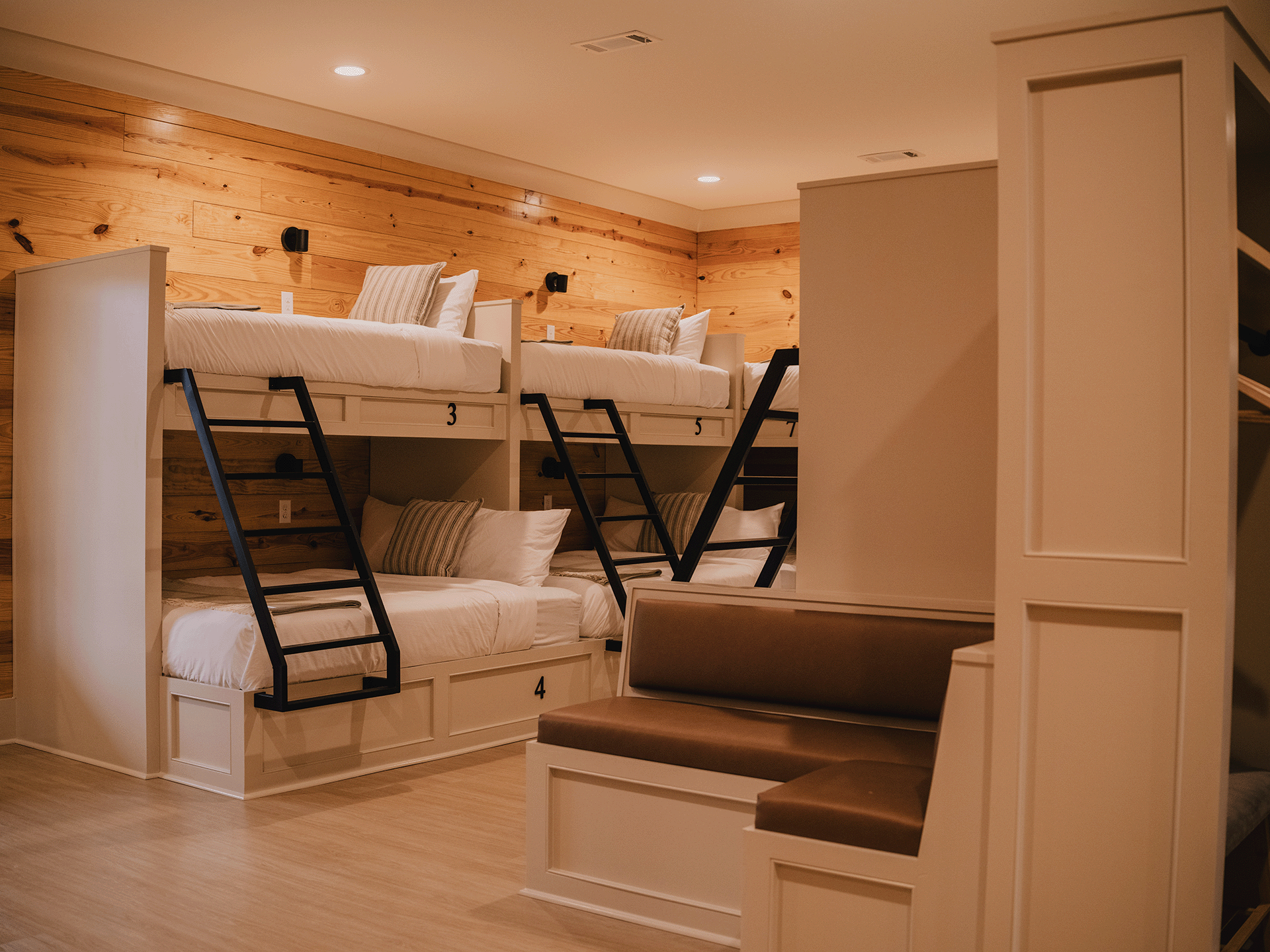 A room with wooden walls featuring white bunk beds with numbered tags, black ladders, and pillows, alongside a seating corner with a cushioned bench.