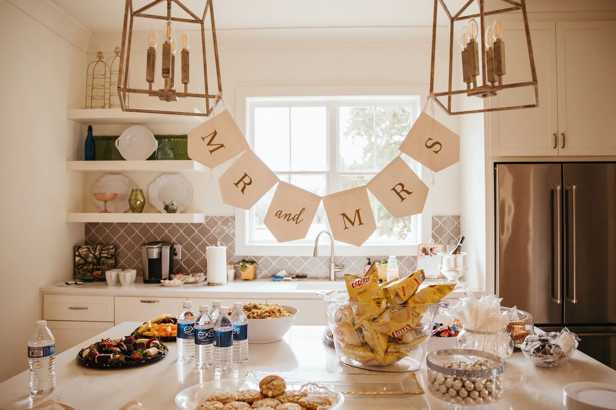 A kitchen decorated for a bridal shower or event with a "Mrs. and Mr" banner hanging above the countertop. The kitchen features white cabinetry, open shelves with dishes and decor, and a window letting in natural light. The countertop has water bottles, snack bowls, cookies, and a large bowl with chips.