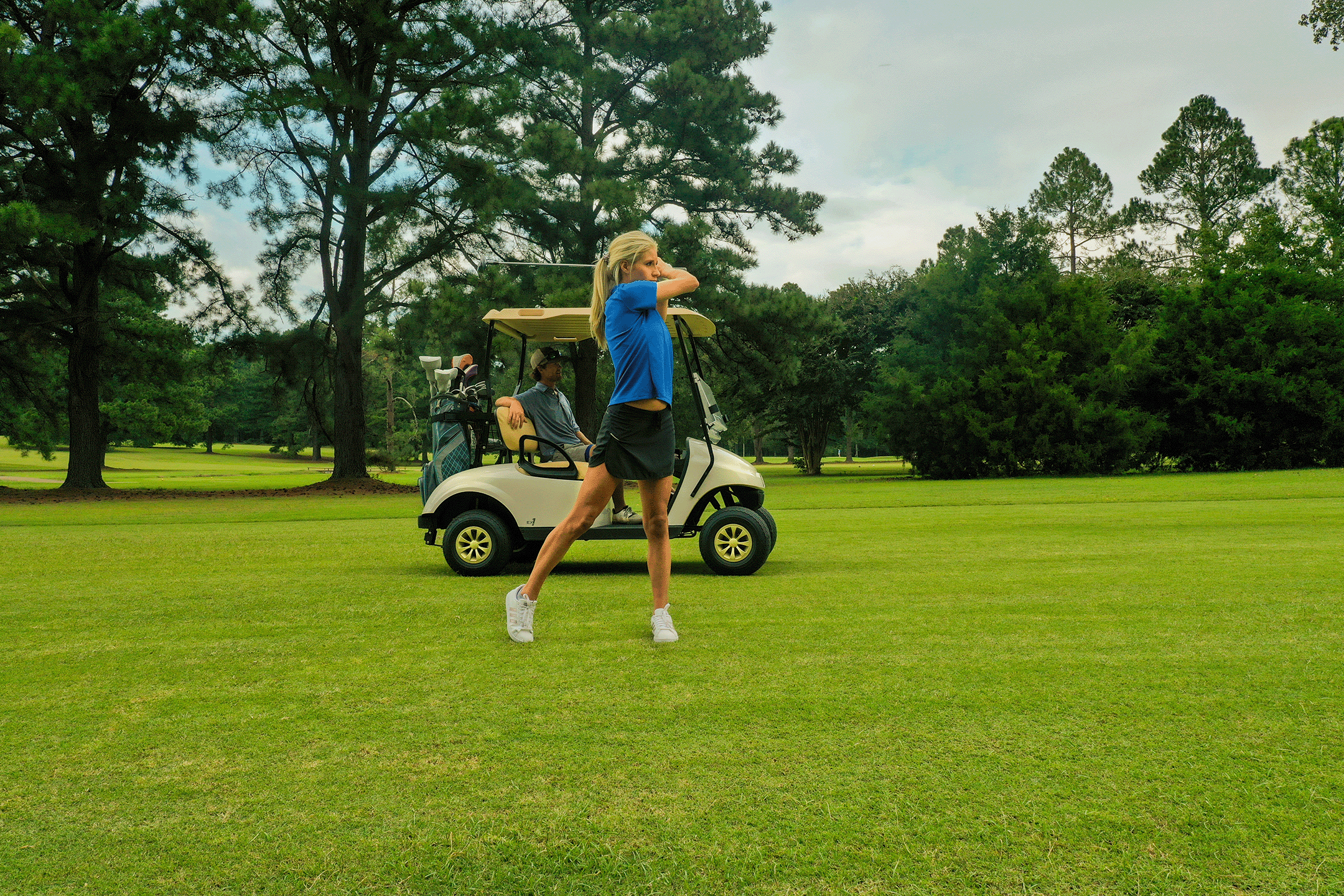 A woman with a ponytail, wearing a blue shirt and black golf skirt, is swinging a golf club on a green golf course. Behind her, there is a golf cart with a man sitting inside, surrounded by trees.