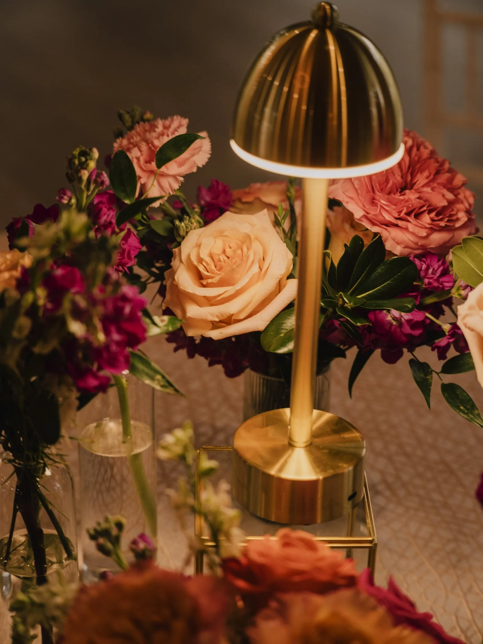 A gold table lamp surrounded by pink and peach roses and other flowers in glass vases.