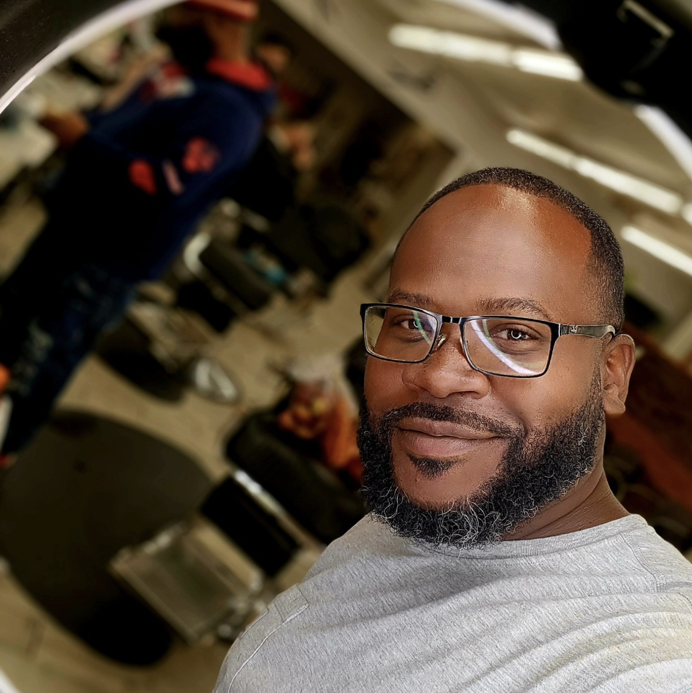 A man with glasses and a beard taking a selfie in a room that appears to be a garage or repair shop, with a person in a navy jacket in the background near various tires and equipment. The owner of meridian construction