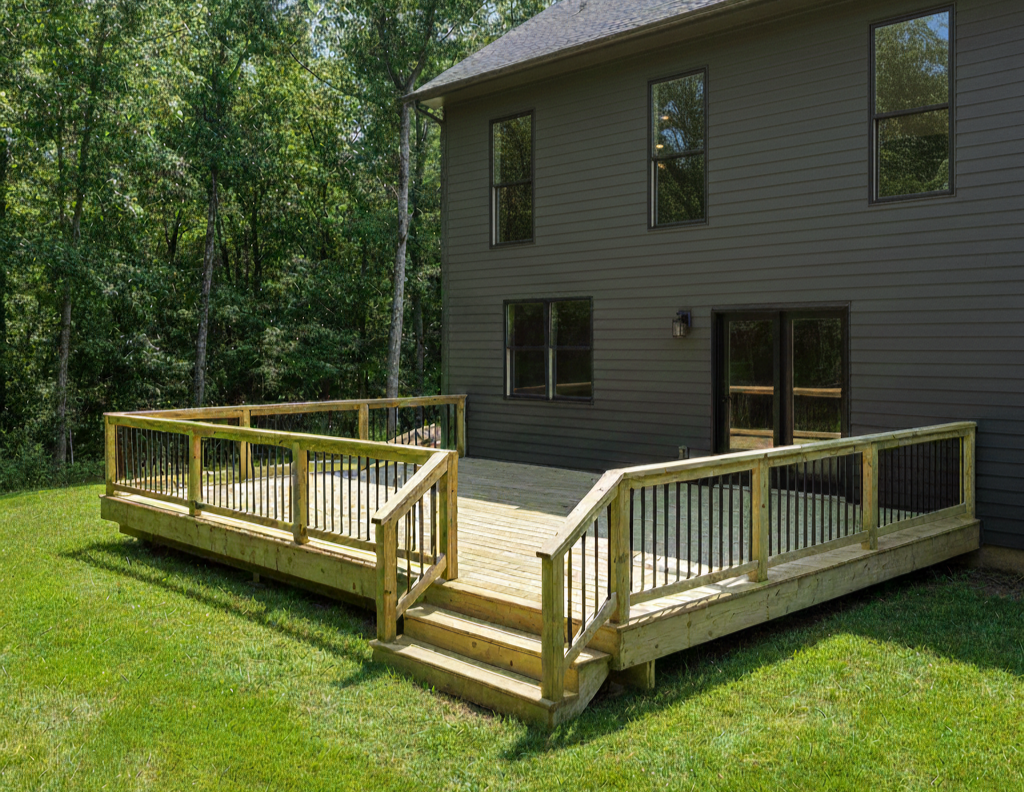 New wooden deck with black metal railings attached to a gray house, surrounded by green grass and trees.