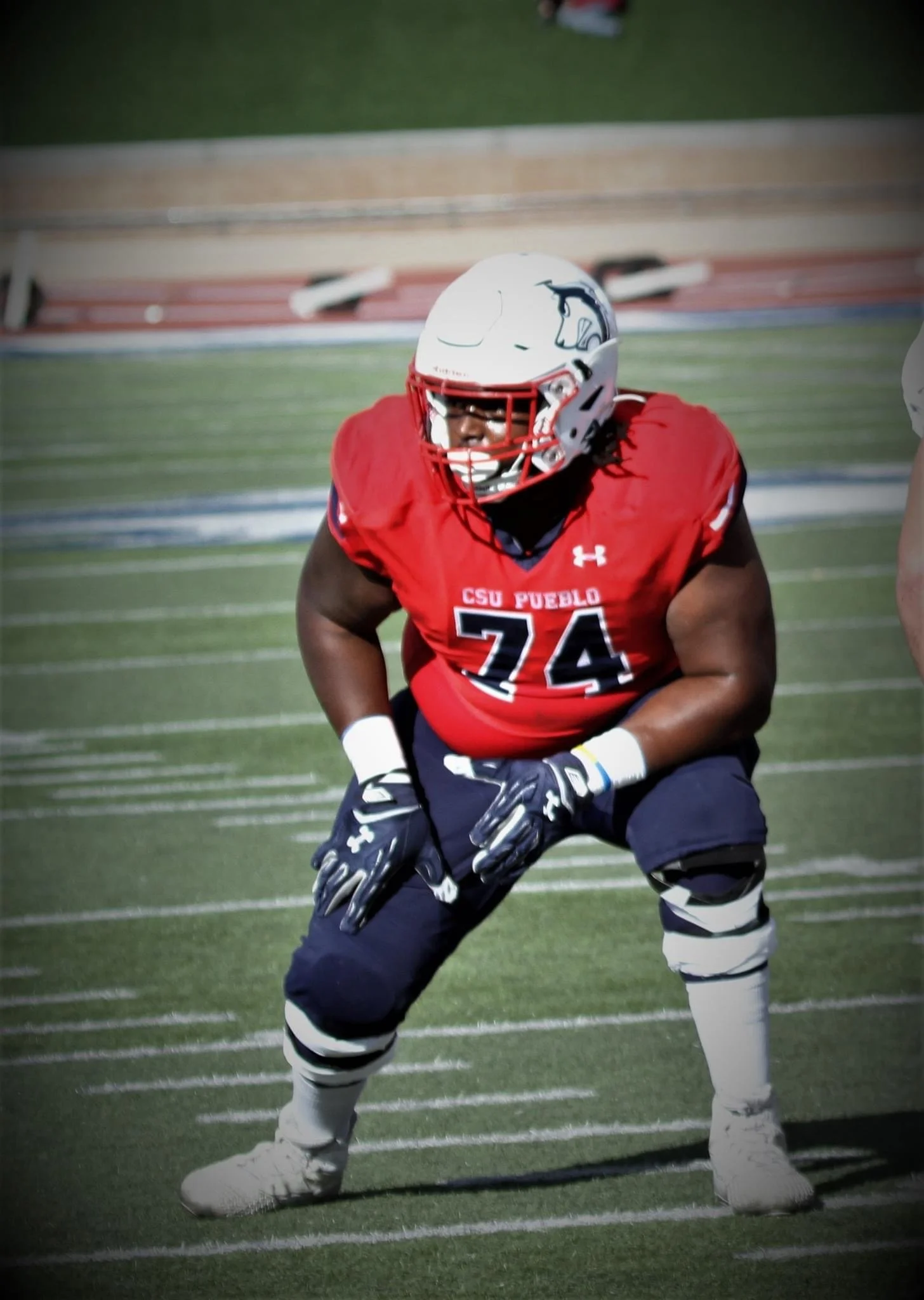 An American football player in a red jersey with the number 74, wearing a white helmet, black gloves, and black pants, crouched on a football field.