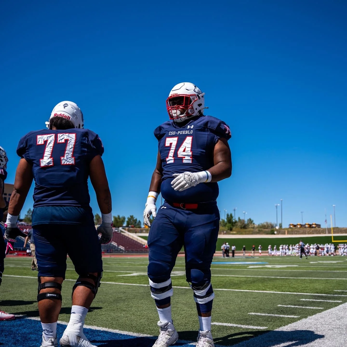 Two American football players on the field wearing navy blue uniforms with red and white details, including helmets, gloves, and knee-high socks, during daytime under a clear blue sky.