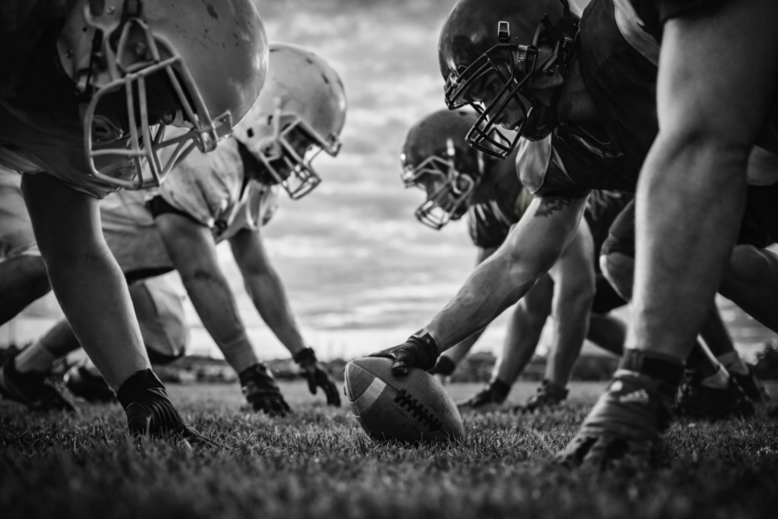 Black and white photo of American football players in formation on the field, preparing for a snap with a football in the center.