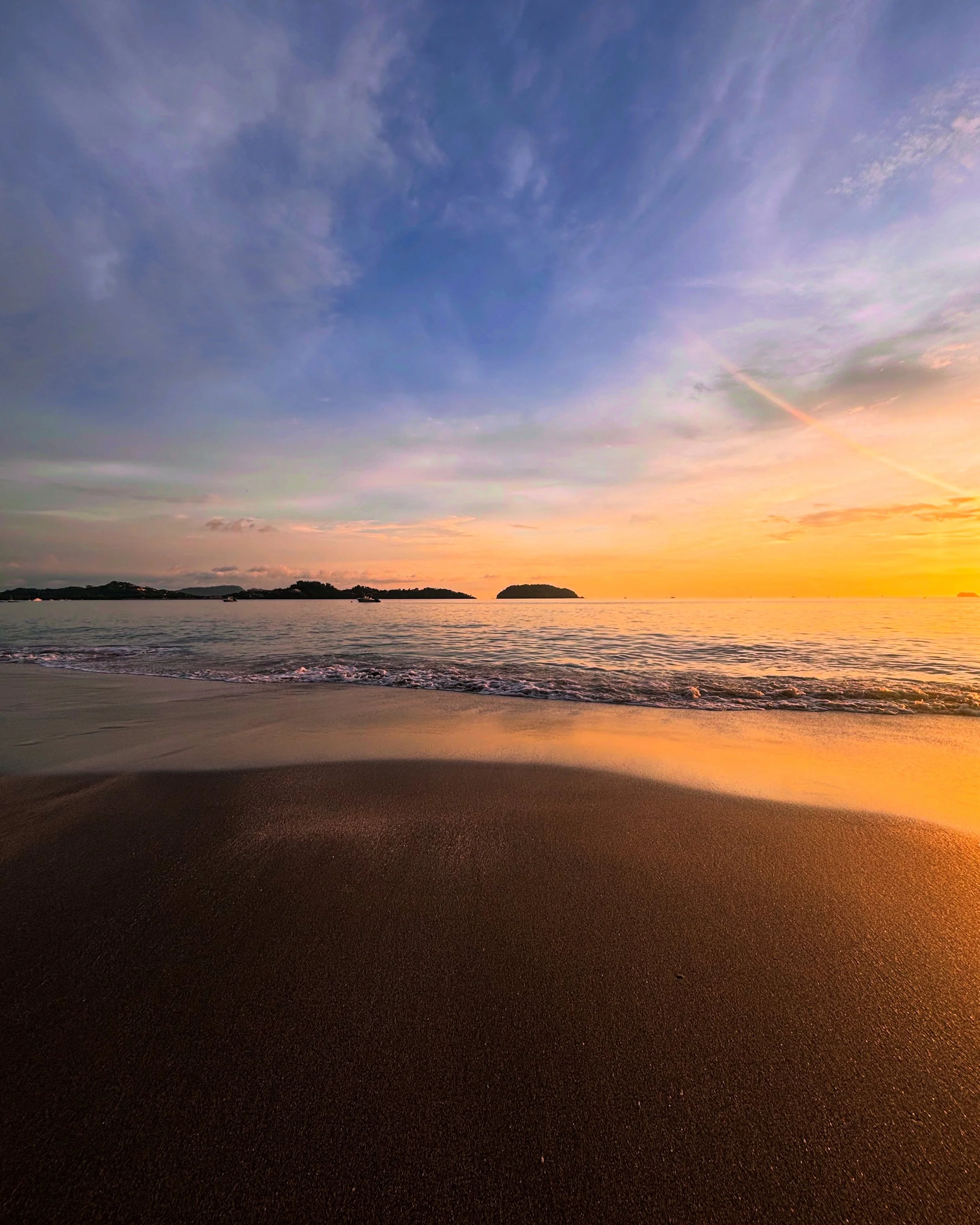 Sunset over a beach with calm waves, sandy shore, and islands in the distance.