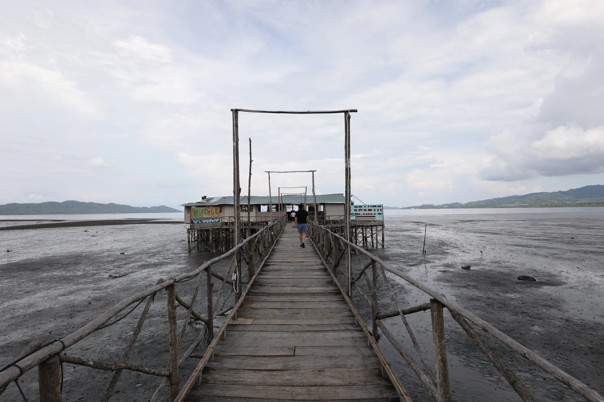 A wooden pier extends over a muddy, low tide area of the ocean toward a stilt house. The house has colorful signs, with one reading 'MUELLE' and another with partially visible text. The sky is overcast with some clouds, and distant forested hills are visible on the horizon.