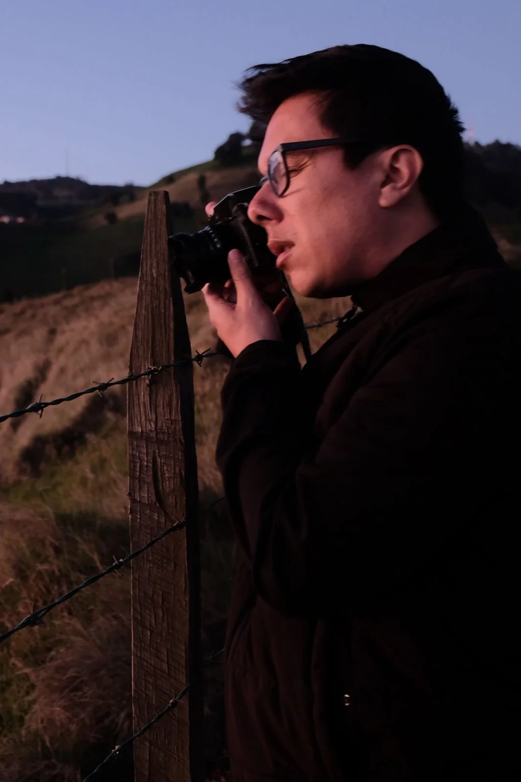 A man with glasses taking a photograph with a camera, standing next to a wooden fence post with barbed wire at dusk or dawn.