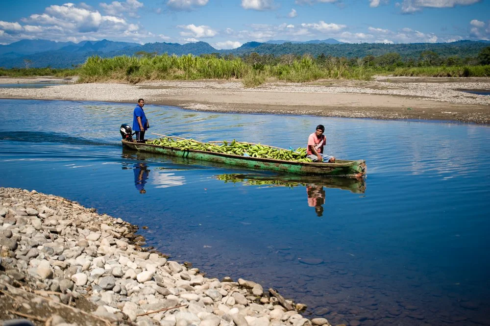 Two men on a small boat filled with bananas crossing a river in a rural area with mountains and cloudy sky in the background.