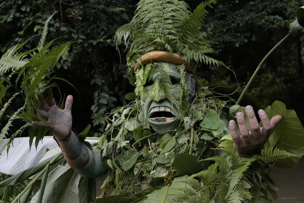 A person dressed as a nature-themed tribal character with a mask that has a wide mouth, wearing a headdress of ferns, surrounded by lush green leaves and plants in a forest setting.