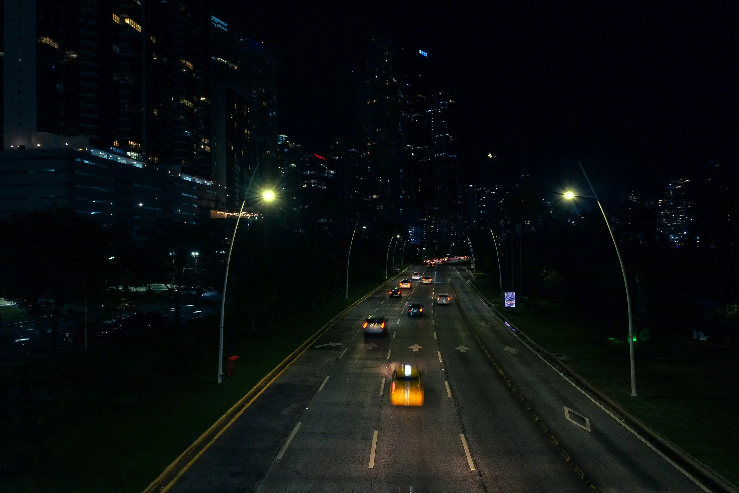 Nighttime city street with moving cars, streetlights, and tall illuminated buildings in the background.