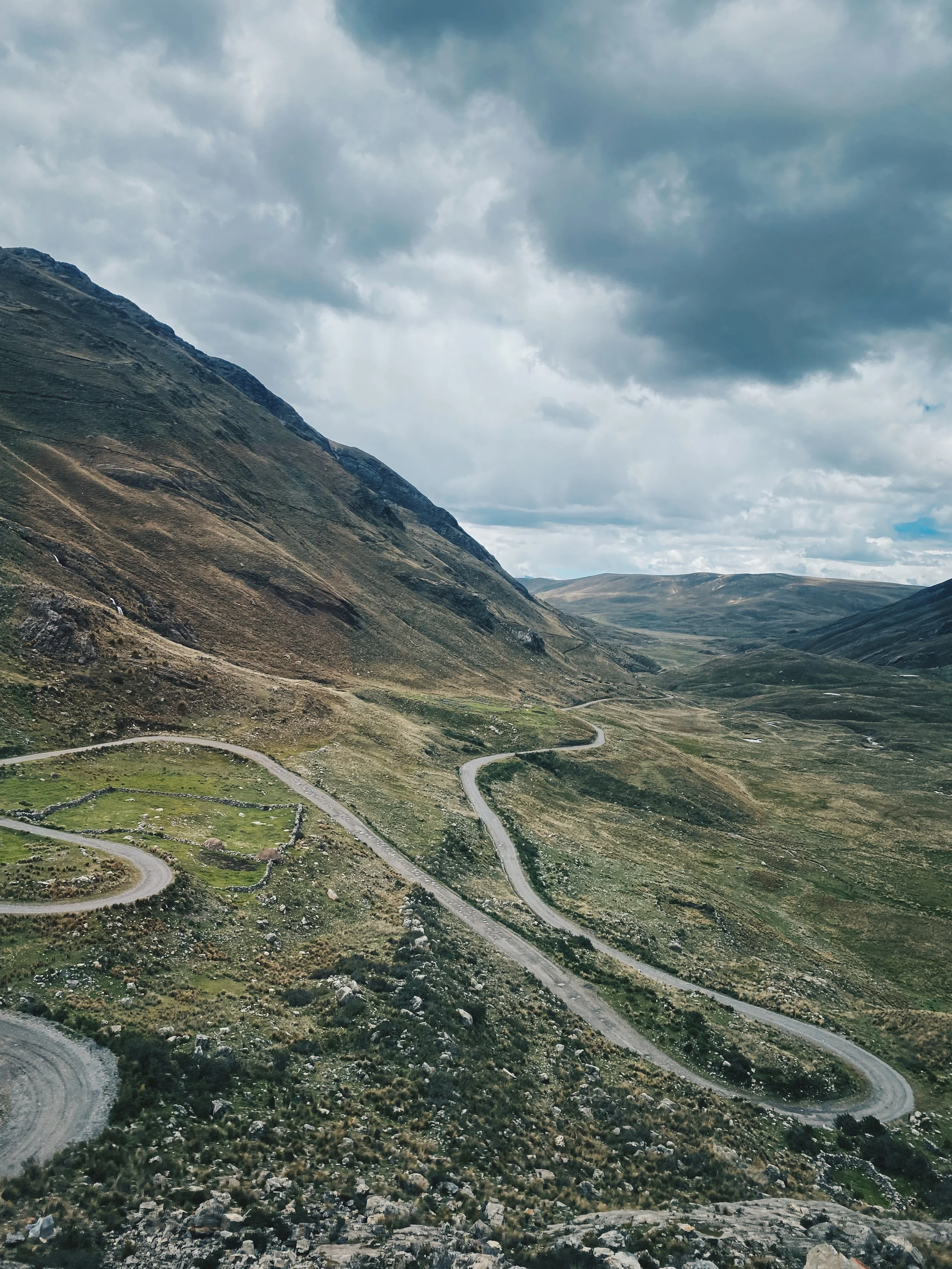 Winding mountain road through green, rocky landscape under cloudy sky.
