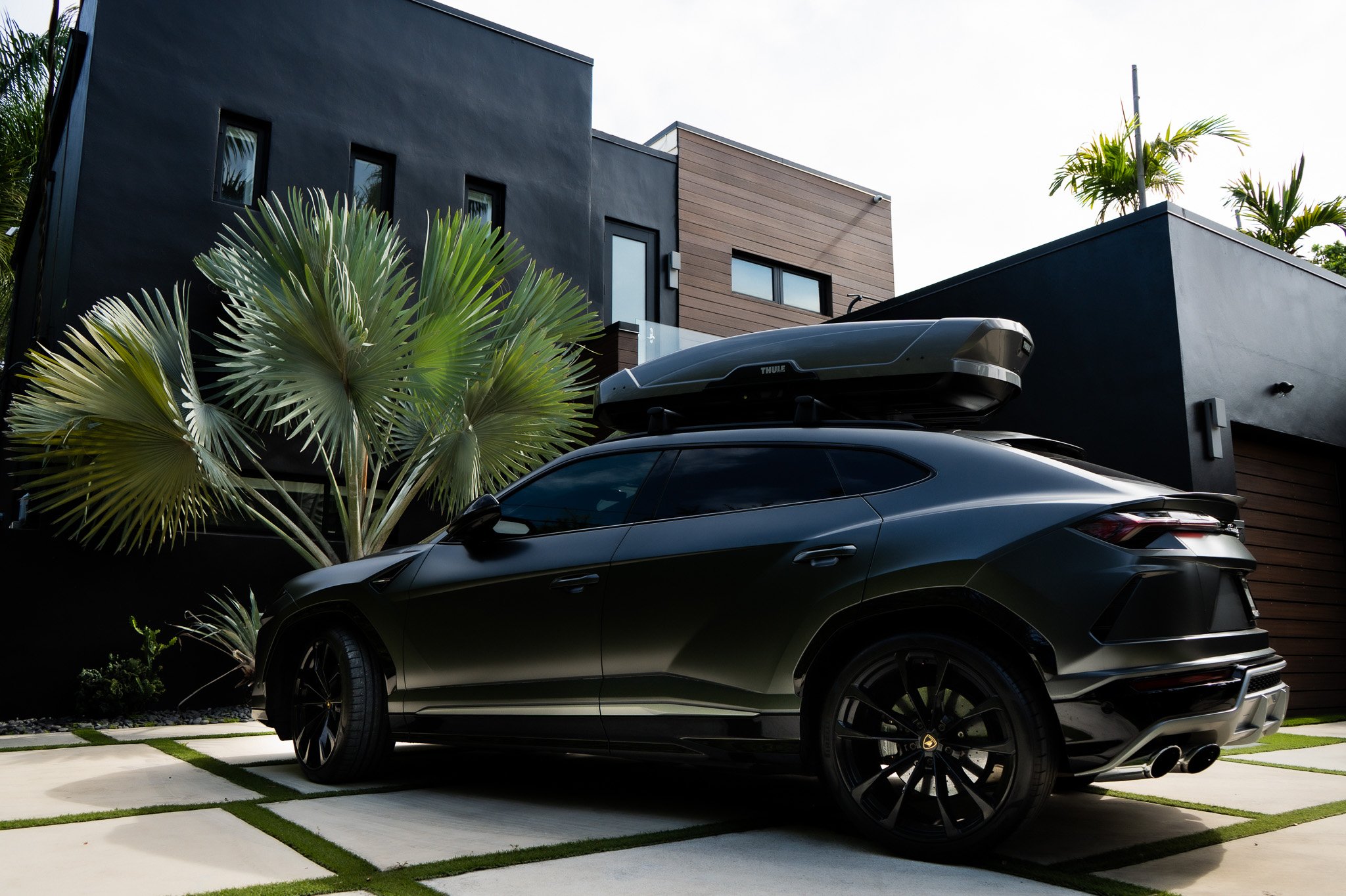 A black luxury car with a roof cargo box parked in front of a modern house with palm trees.