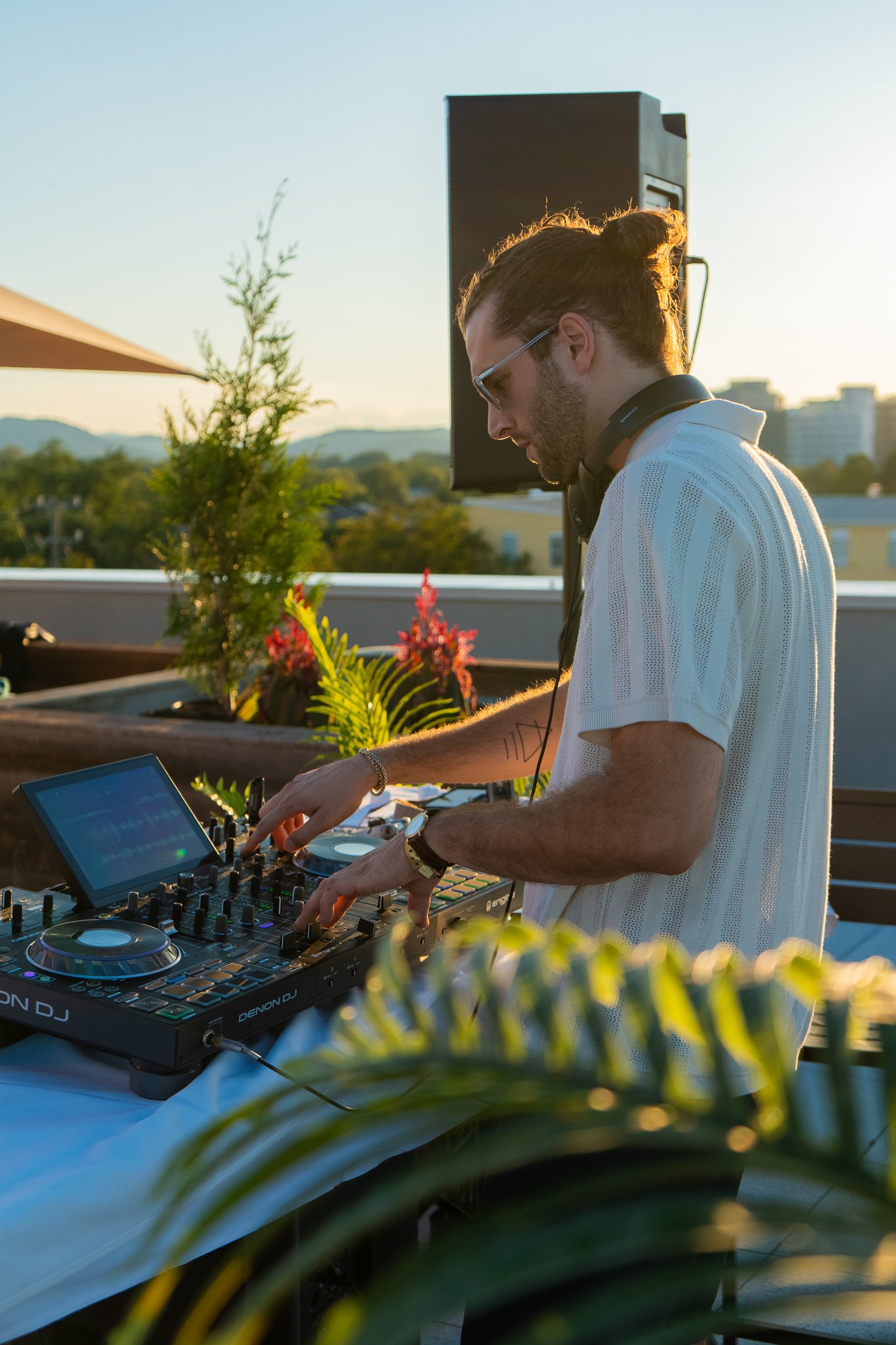 A DJ wearing sunglasses and a white shirt is mixing music on a DJ controller at an outdoor event during sunset. There are plants and city buildings in the background.