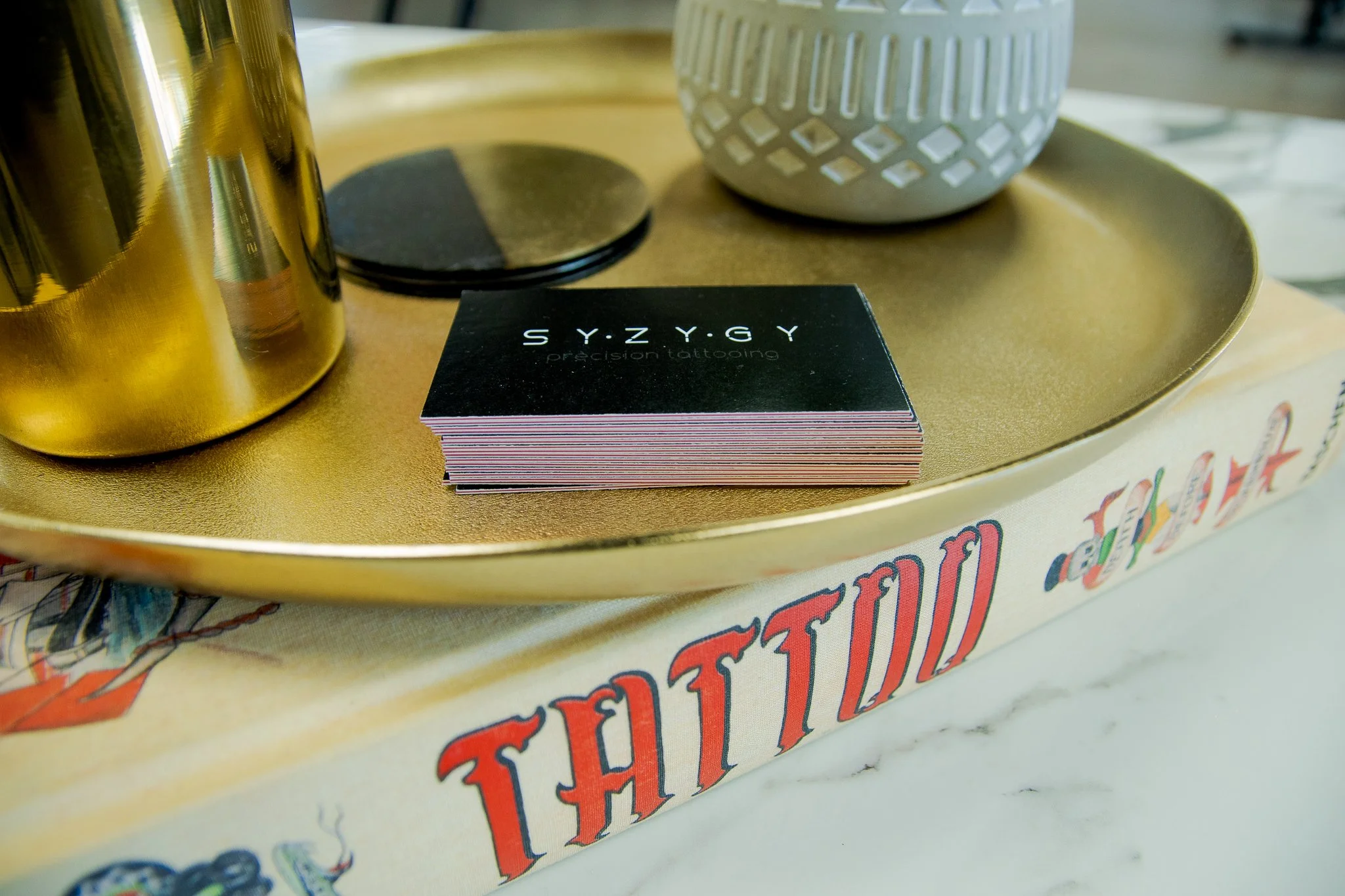 Close-up of a black business card with white text reading 'S Y. Z Y. G Y precision tattooing,' placed on a gold-colored tray along with a gold tumbler, a white container, and a stack of circular black coasters on a table with colorful artwork and tex