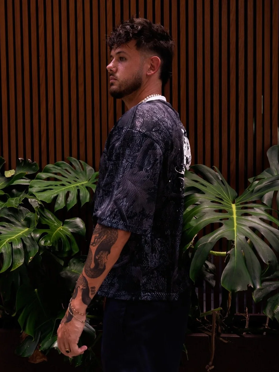 A man with dark curly hair and a beard, wearing a black patterned shirt, standing sideways in front of a wooden wall and surrounded by large green leafy plants.