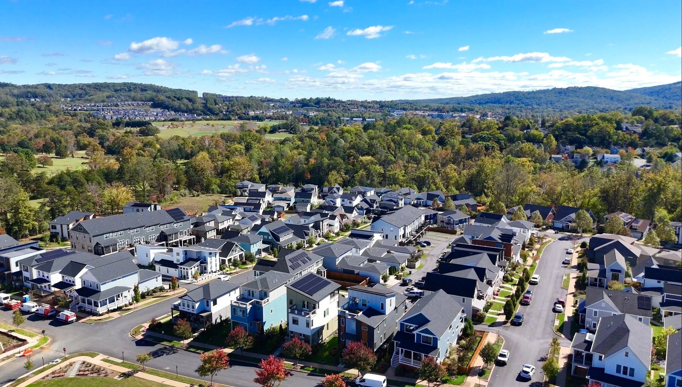 Aerial view of a suburban neighborhood with new houses, some with solar panels, streets, parked cars, and trees, surrounded by greenery and rolling hills under a partly cloudy sky.