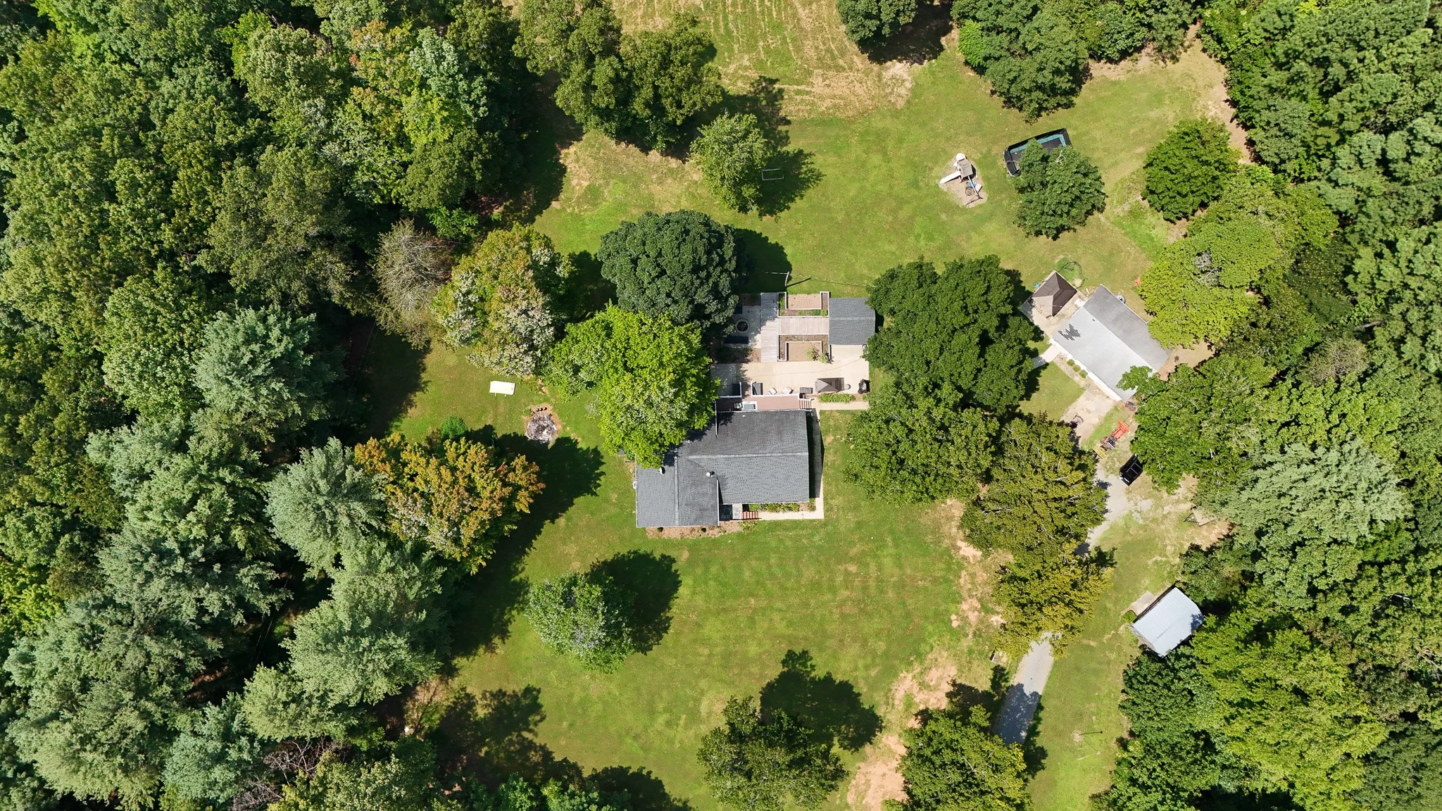 Aerial view of a house surrounded by trees, greenery, and open lawn space, with some small structures nearby.