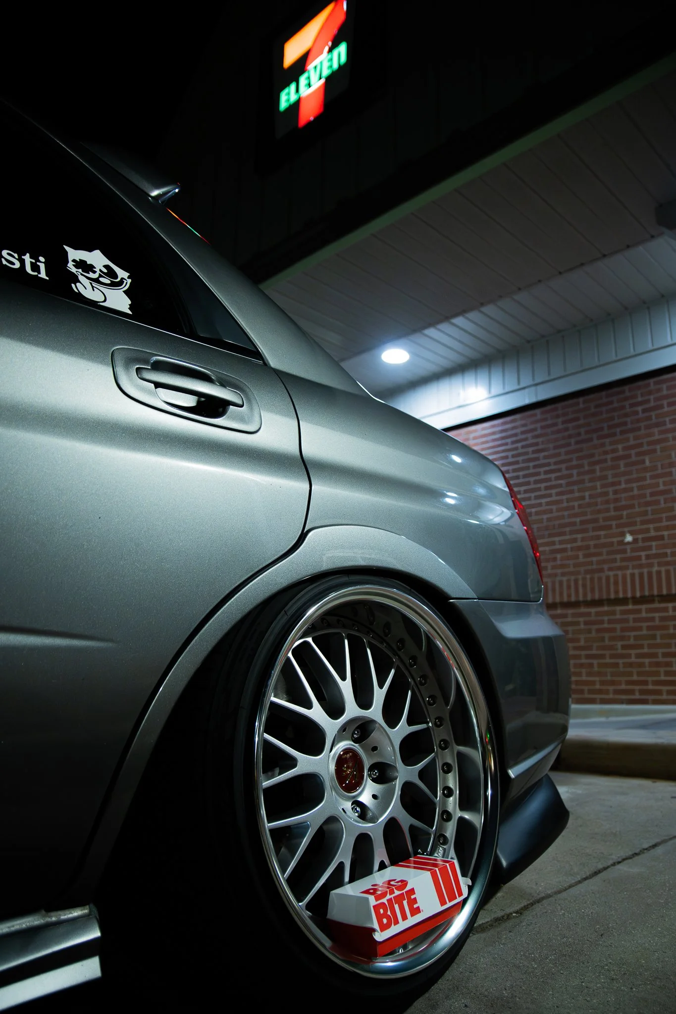 Close-up of the rear side of a gray car parked outside a 7-Eleven convenience store at night, with custom wheels and a box of 7-Eleven Big Bites snack on the ground near the wheel.