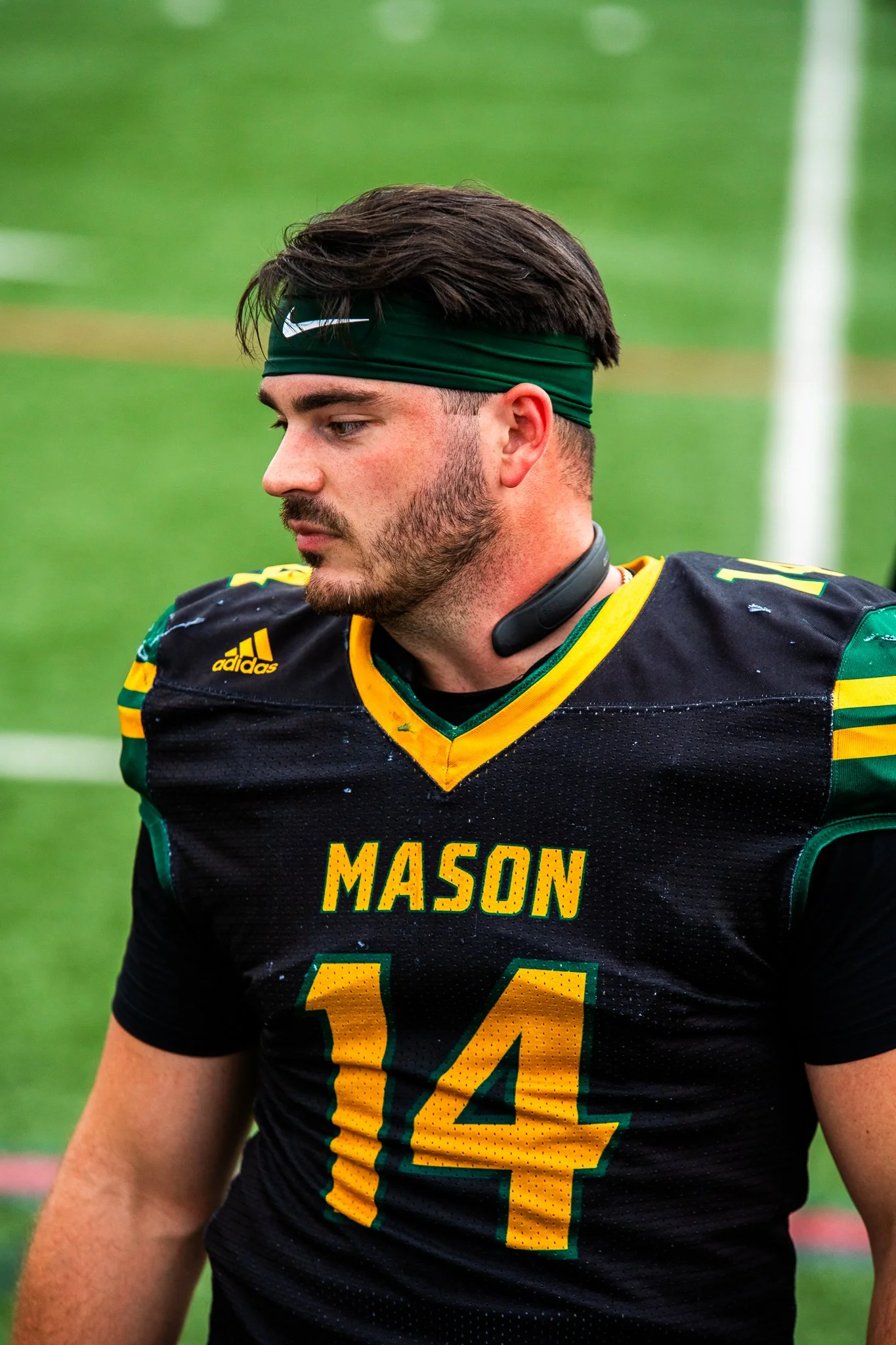 A young male football player wearing a black and yellow 'Mason' jersey with the number 14, a green headband, and a neck fan, stands on a football field with a focused expression.