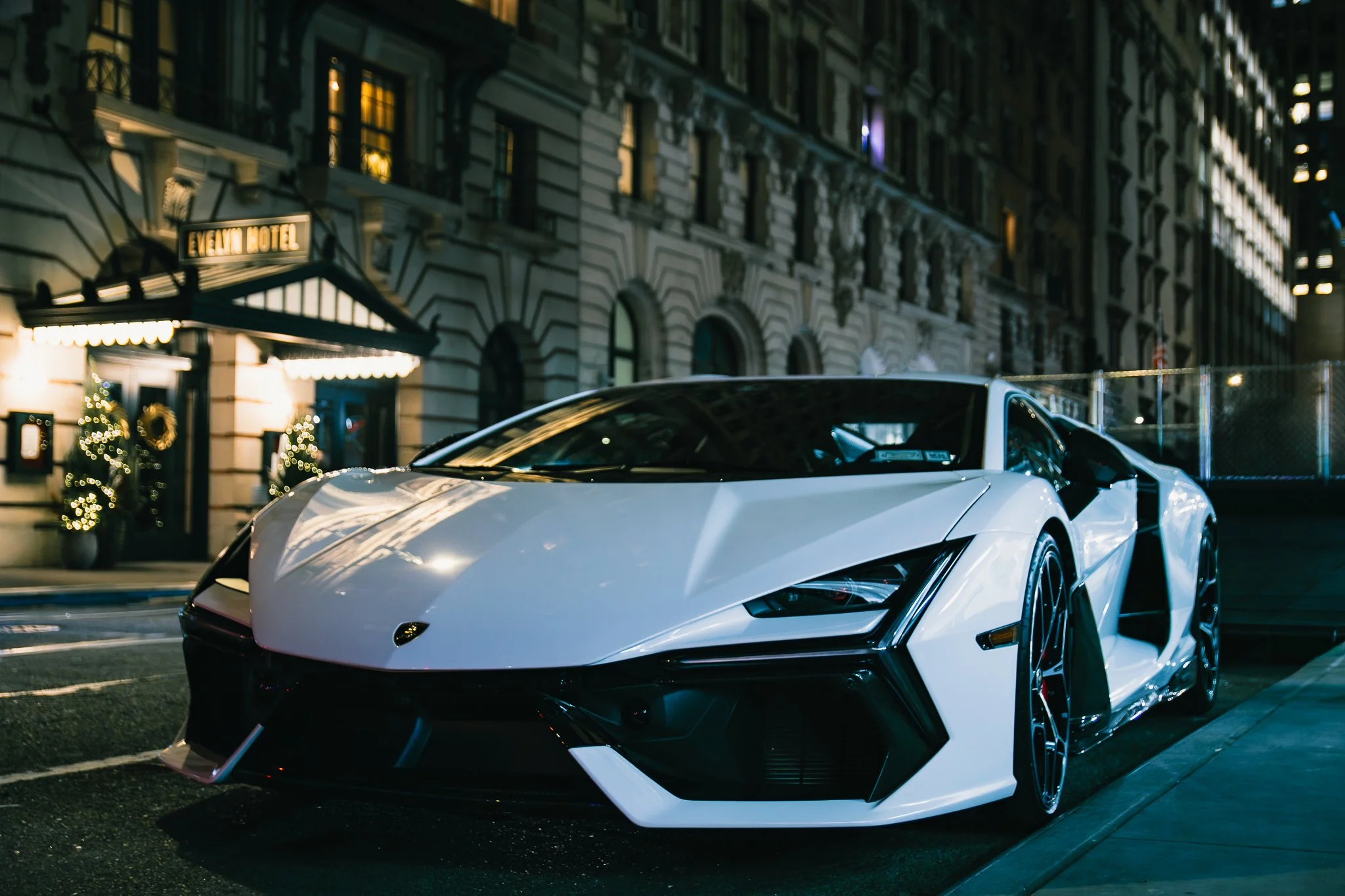 A white luxury sports car parked on a city street at night, with the hotel in the background decorated with Christmas lights.