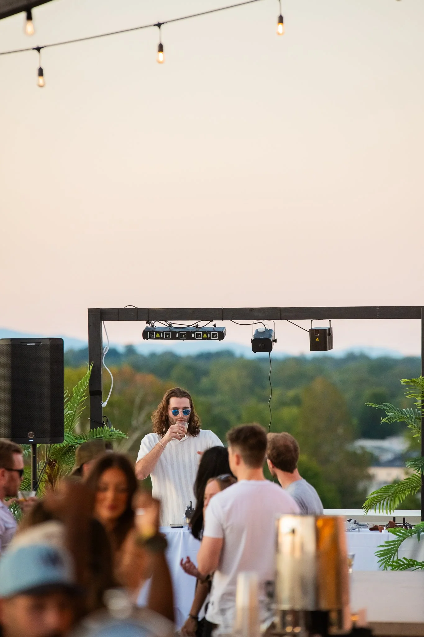 A man with long hair and sunglasses is drinking from a glass at an outdoor party, with people and trees in the background.