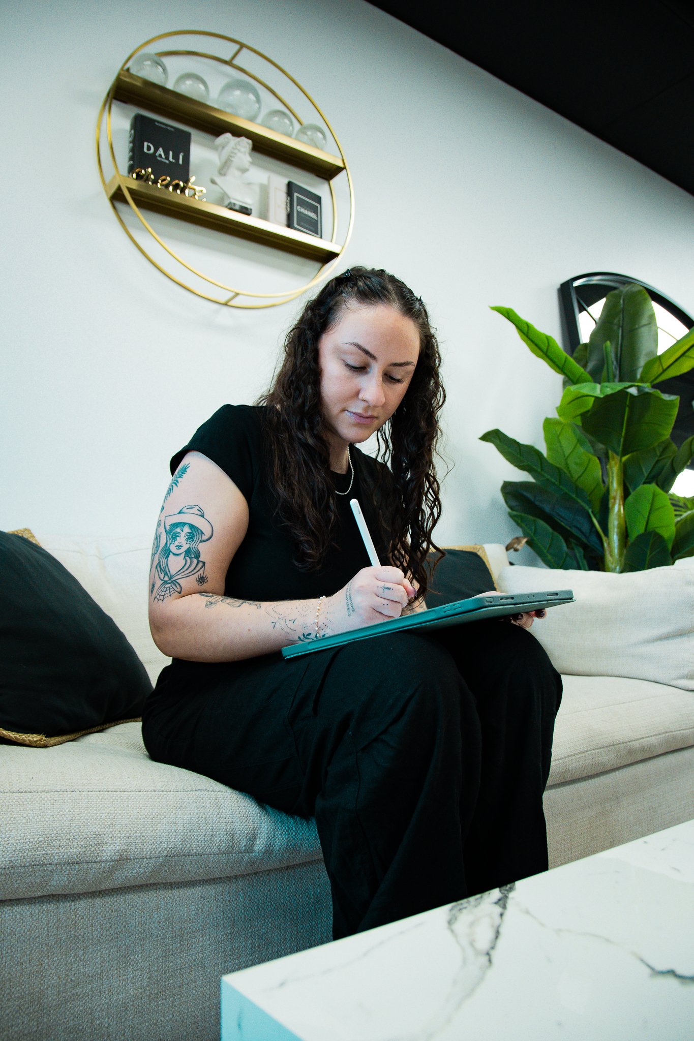 A woman with curly hair, tattoos on her left arm, dressed in black, sitting on a beige sofa with black pillows, writing on a digital tablet with a stylus. There is a large houseplant to her right and a gold circular wall shelf with decorative items a