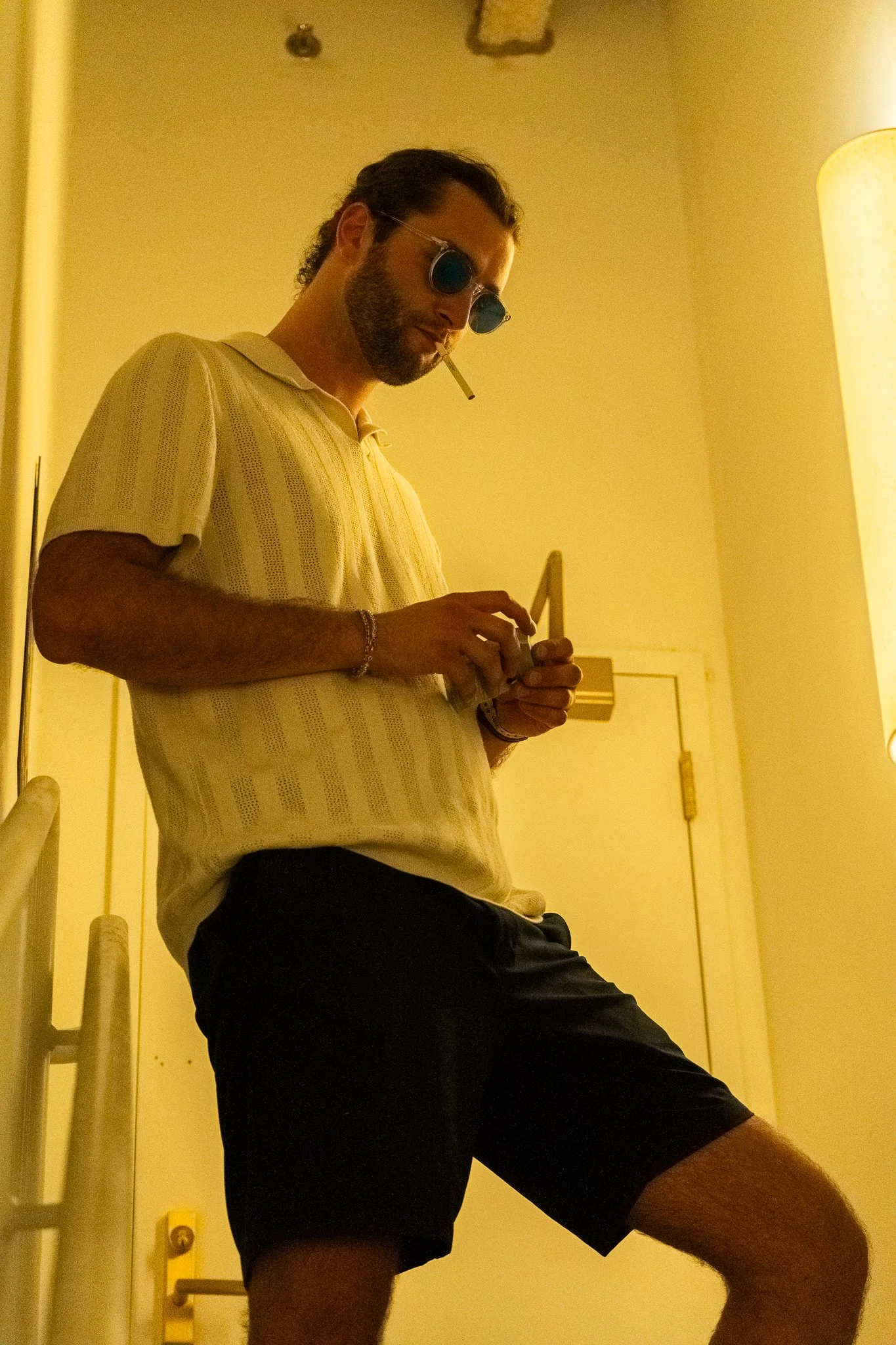 A man with sunglasses and a cigarette in his mouth, dressed in a yellow shirt and black shorts, standing in a dimly lit room, looking at his phone.