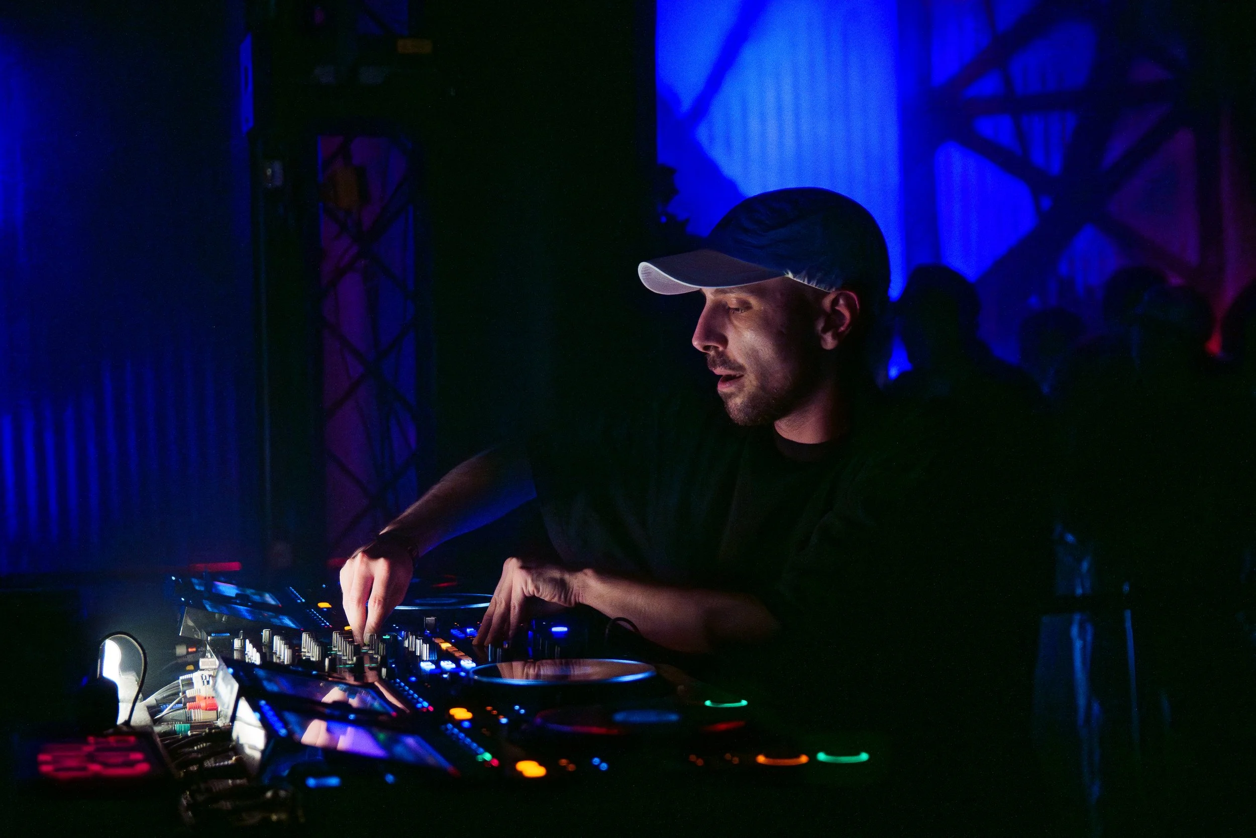 A DJ wearing a cap performs at a nightclub with blue and red lighting, using a mixer and turntables.