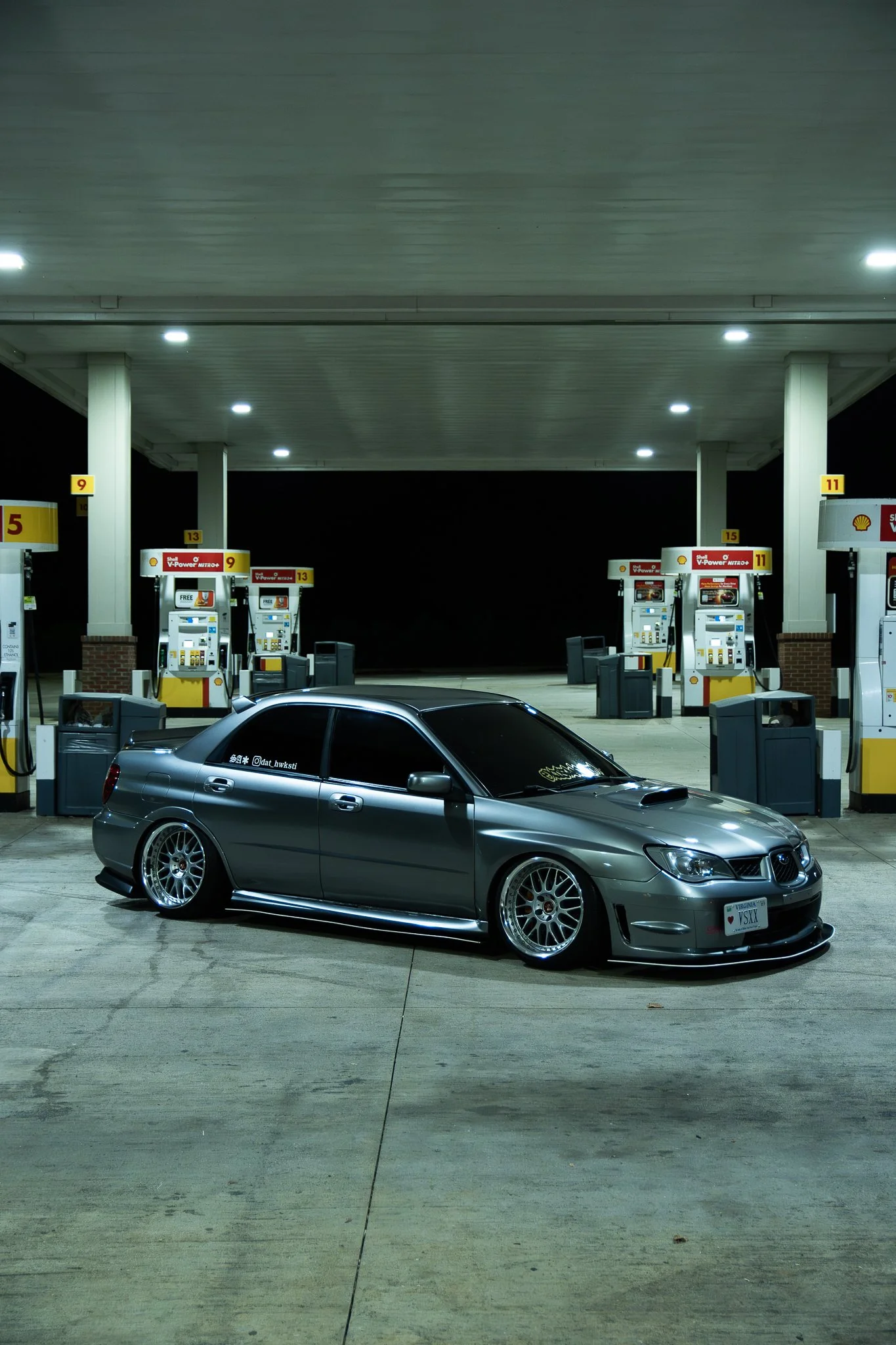 A silver modified Subaru sedan with aftermarket wheels parked at a Shell gas station at night.