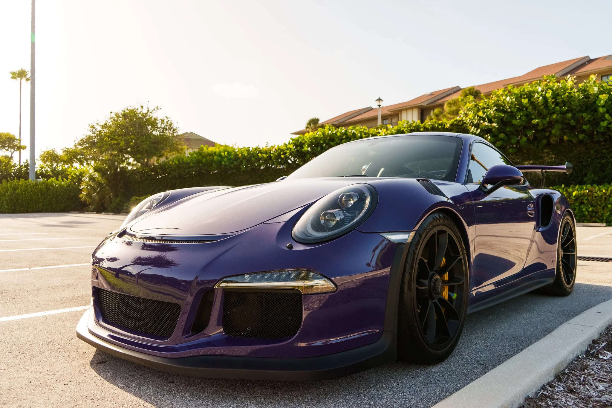 A purple Porsche sports car parked in an outdoor parking lot with trees, shrubs, and buildings in the background.