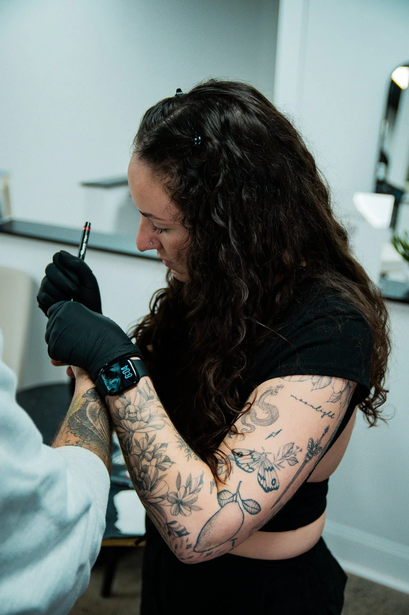 A woman with curly hair tattooed arm getting a tattoo done by a tattoo artist.