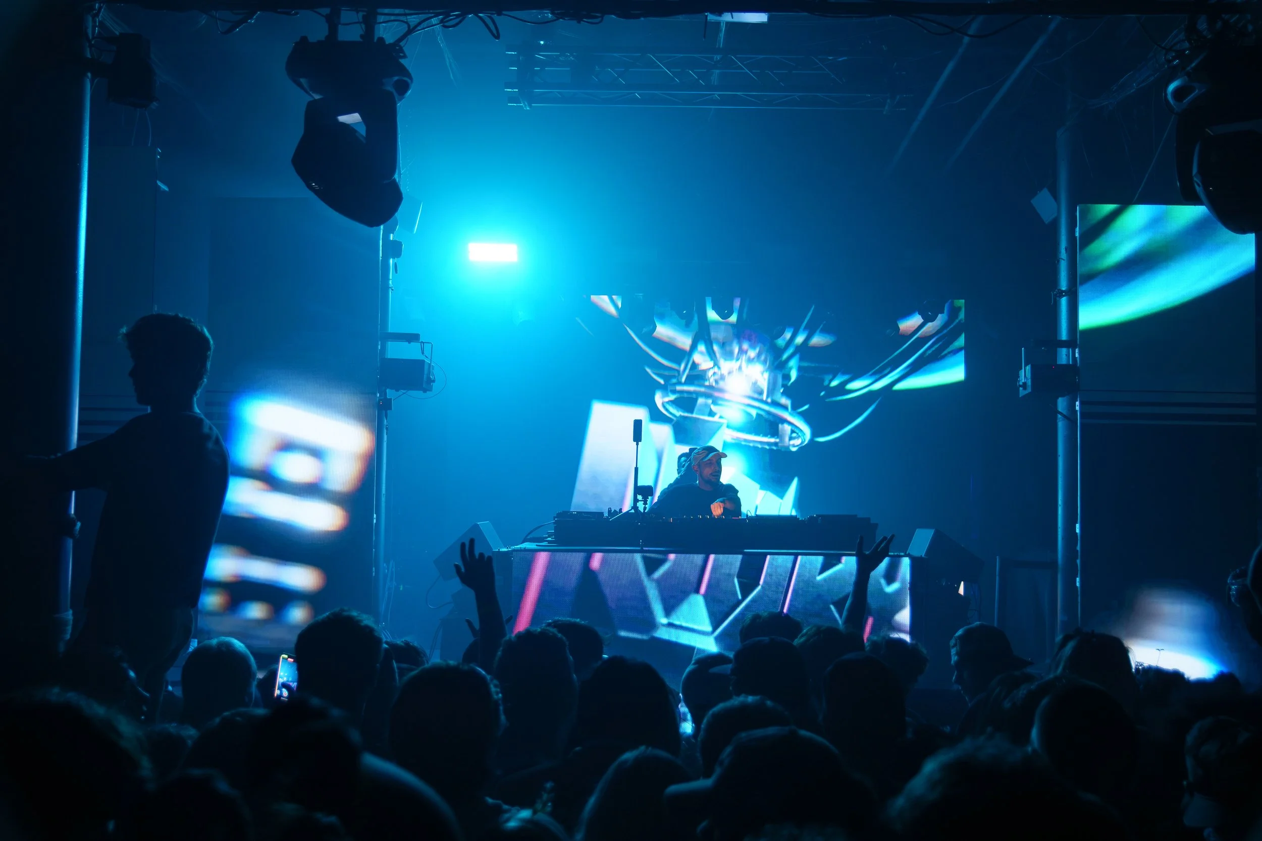 A DJ performs at a concert with a large crowd, blue lighting, and digital screens displaying colorful abstract designs.