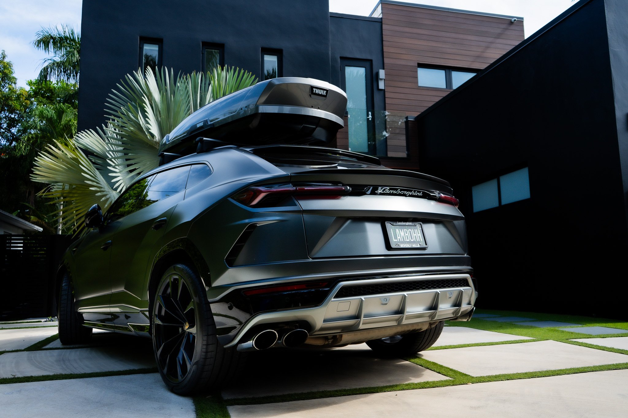 A black Lamborghini SUV parked in a modern driveway with greenery and a contemporary house in the background, with a car roof cargo box on top.