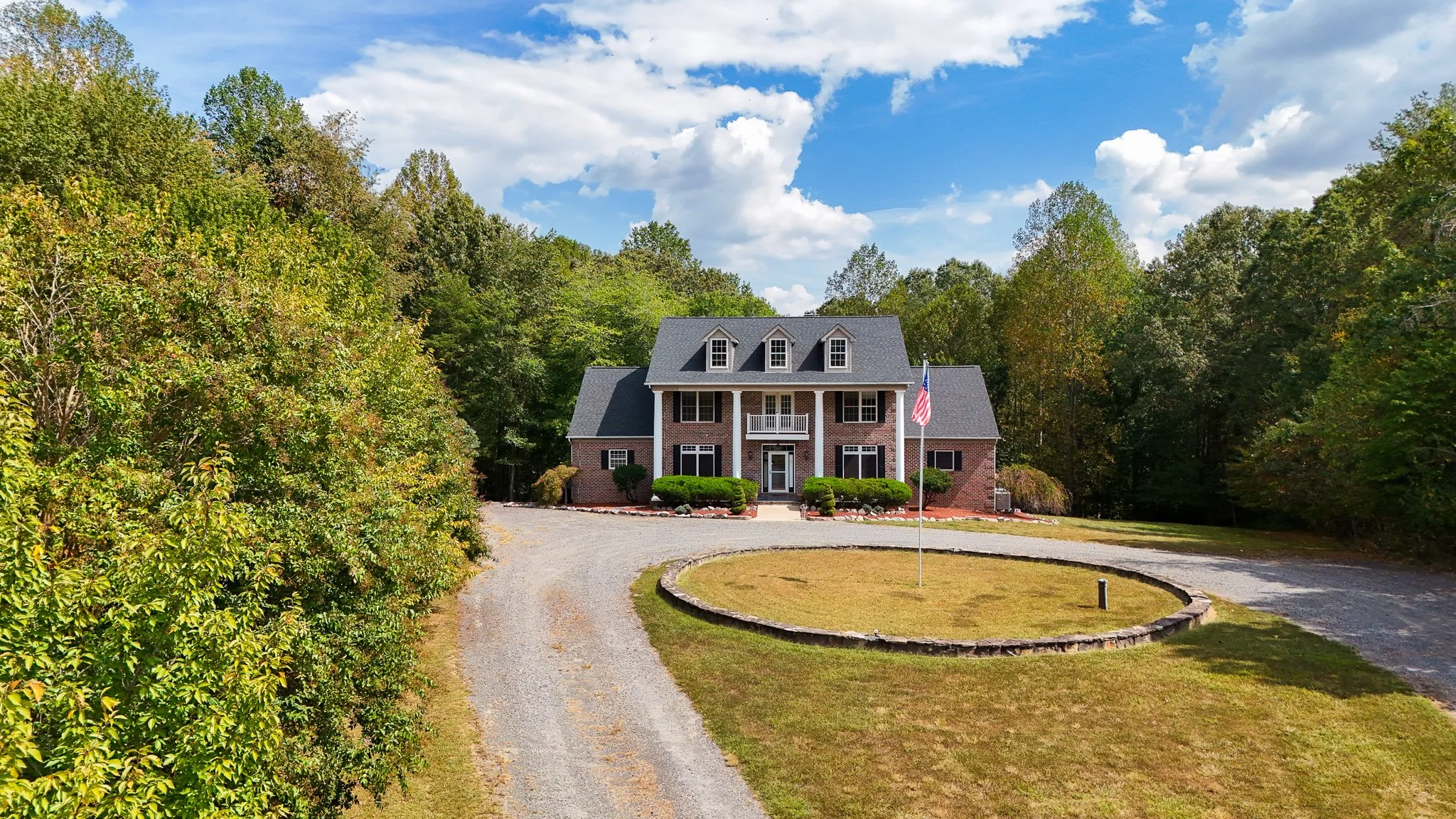 A large brick house with a gray roof and three dormer windows, surrounded by trees. A curved driveway leads to the house, with a circular grassy area and an American flag in the front yard.