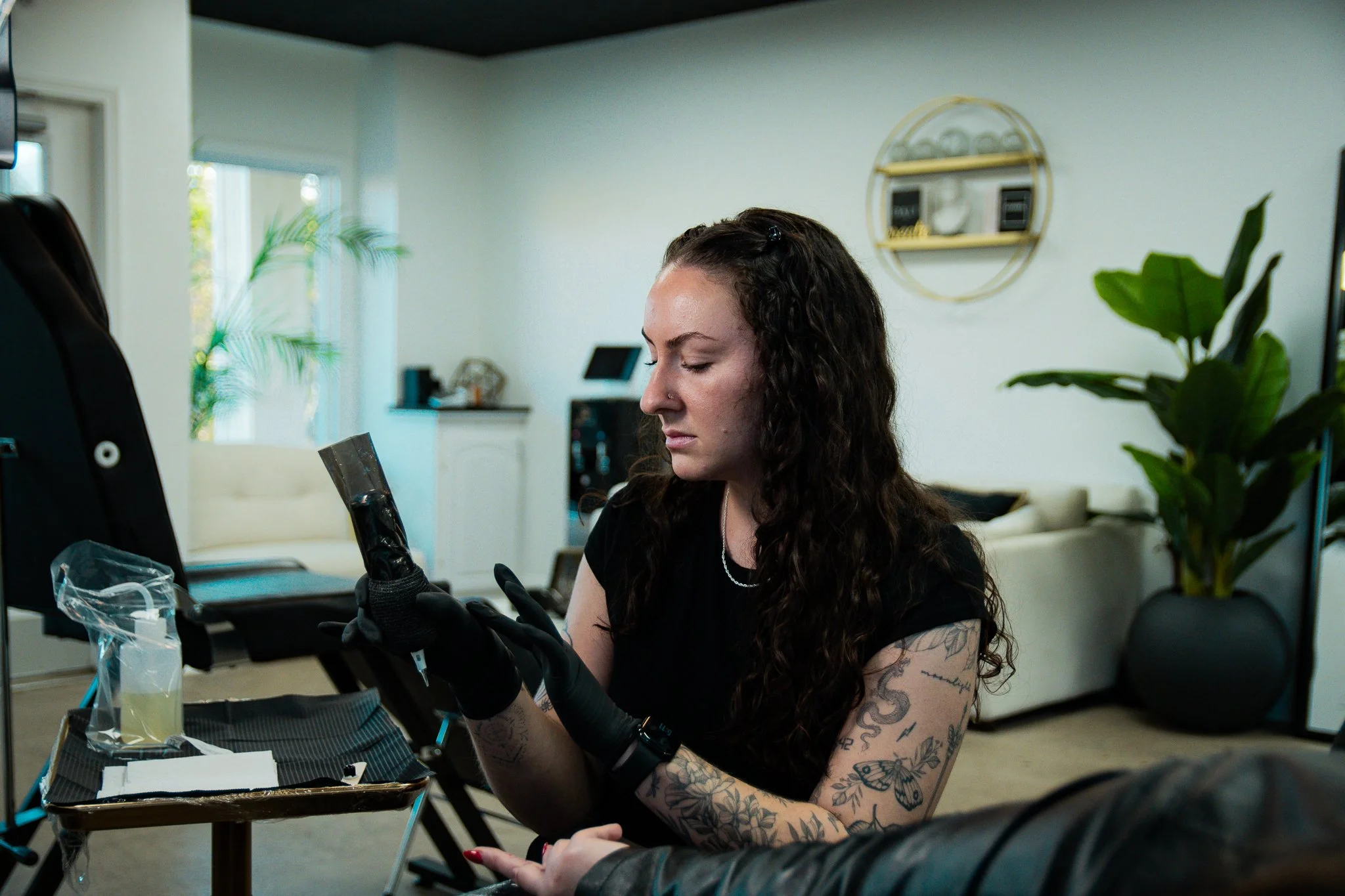 A woman with long tattoos on her arms and dark curly hair, wearing black gloves, is looking at a tattoo design in a home tattoo studio.