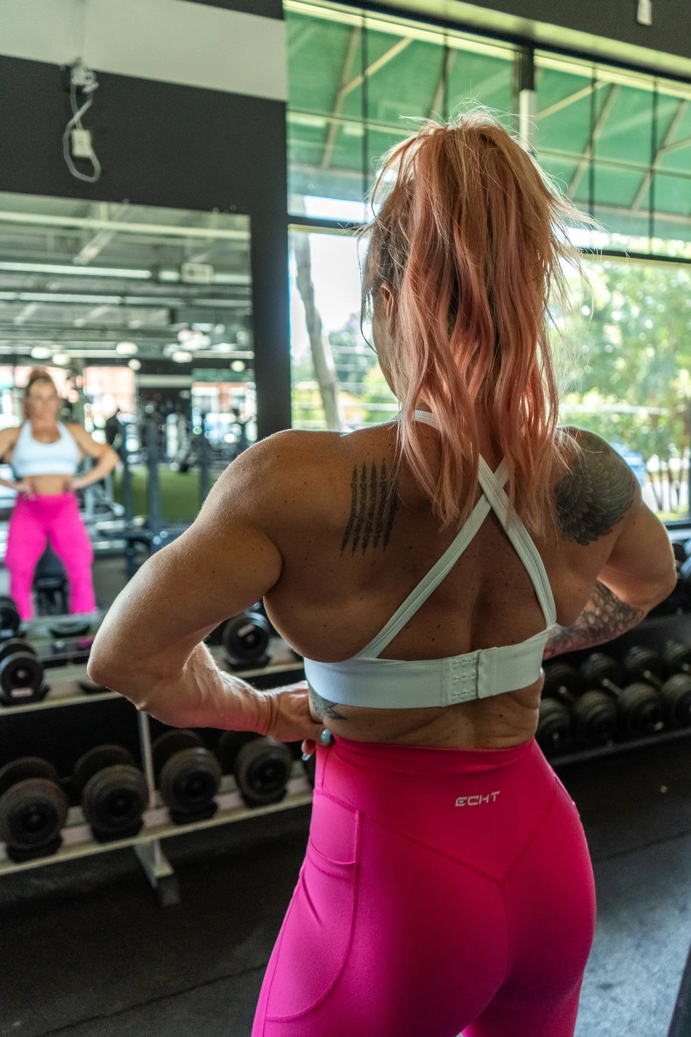 A woman with red hair in a gym, facing away, wearing a white sports bra and pink leggings, with dumbbells and mirrors behind her.