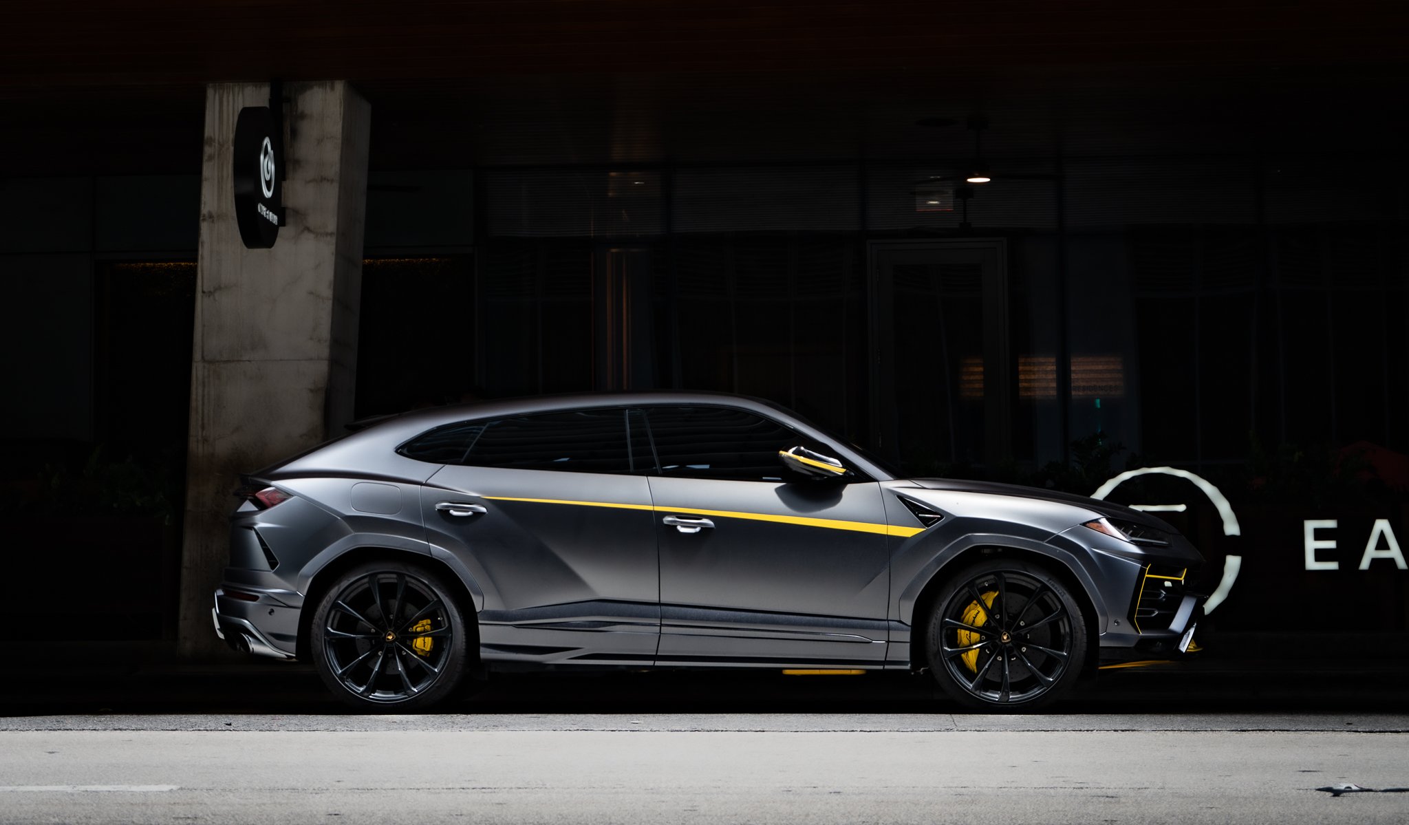 A sleek, dark gray luxury SUV with yellow accents, parked outside a modern building at night.