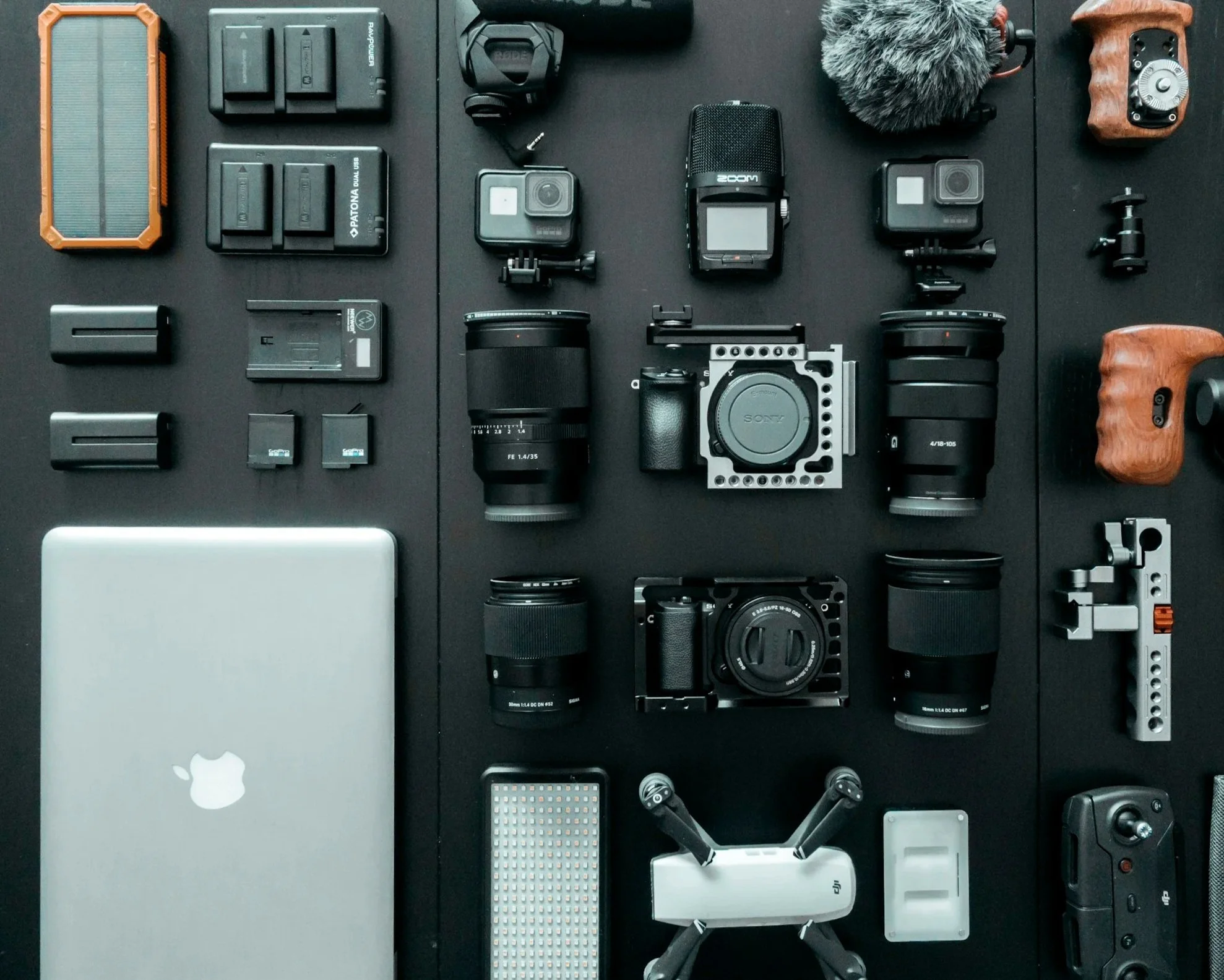 An organized flat lay of various photography and tech equipment on a black surface, including cameras, lenses, batteries, a drone, a laptop, a microphone, and accessories.