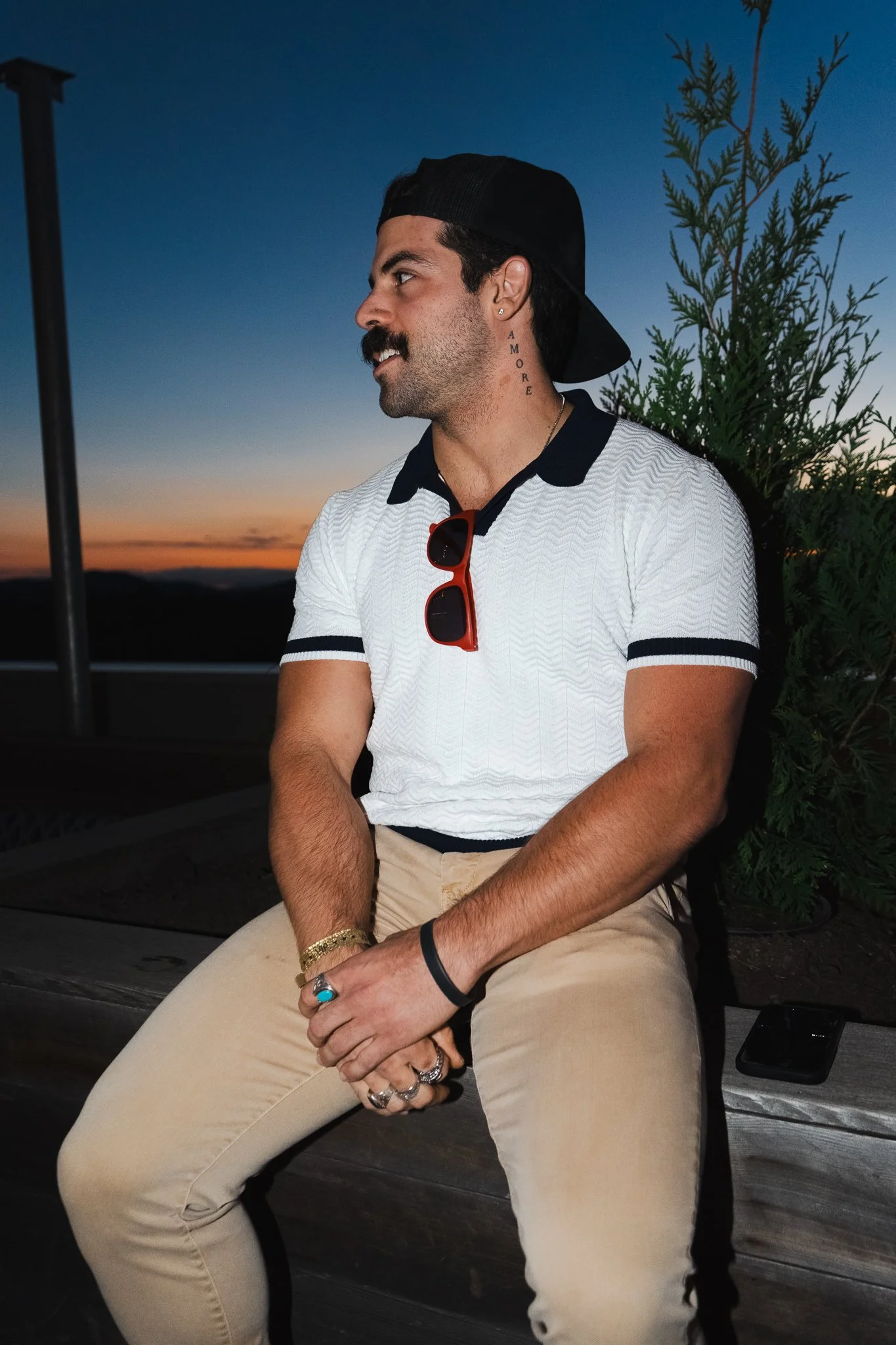Man with dark hair and a moustache, wearing a white polo shirt with black trim, beige pants, a black backward cap, and red sunglasses hanging from his collar, sitting outdoors at sunset with a green plant behind him.