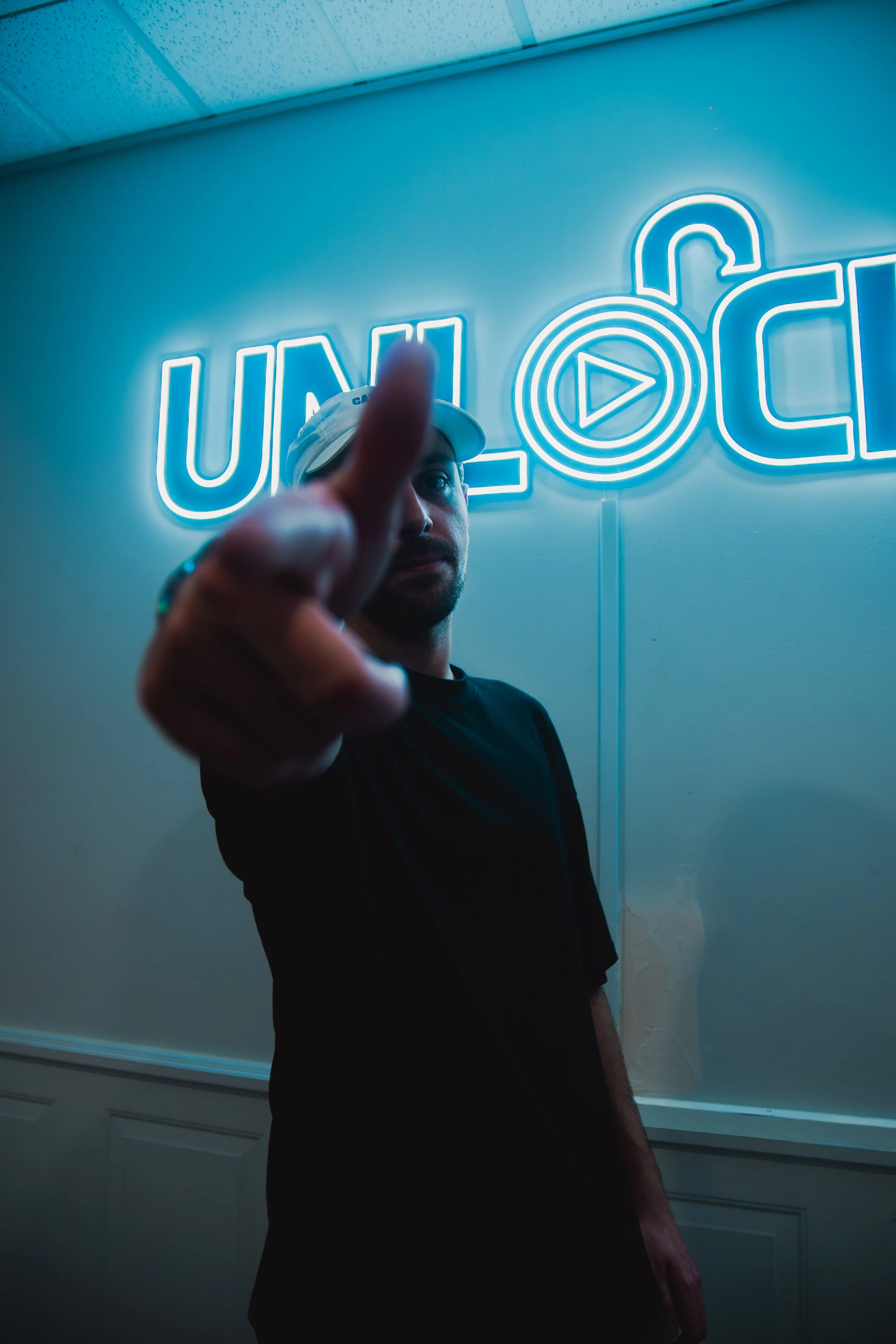 A man in a black shirt and white cap standing in front of a neon sign with the words 'ULLA C' and a lock symbol with a play button in the middle. The man is reaching out with his finger towards the camera.