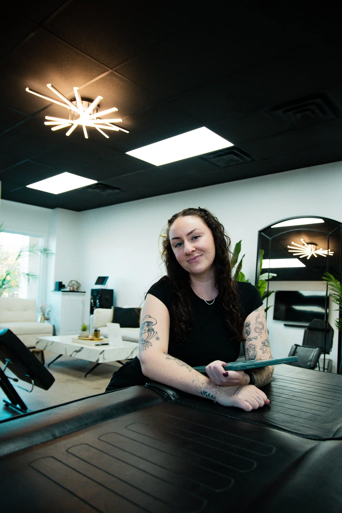 A woman with tattoos on her arms sits behind a counter in a modern, well-lit lounge or reception area, smiling at the camera.